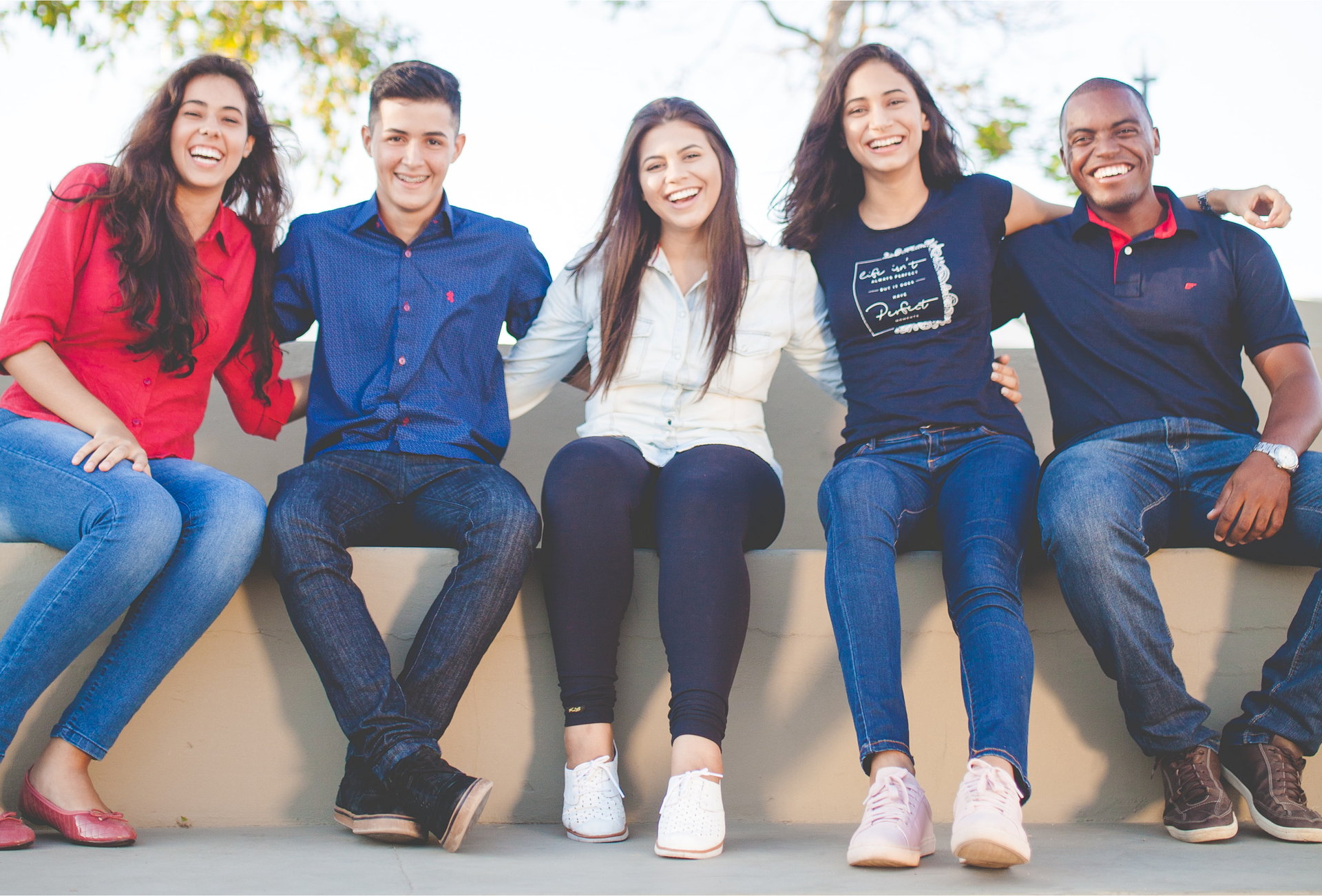 Zdjęcie przedstawia pięcioro młodych, roześmianych osób, pozujących do zdjęcia. Od lewej siedzą: młoda, roześmiana dziewczyna z długimi włosami, następnie siedzi uśmiechnięty chłopak z krótkim włosami, dalej siedzą obok siebie dwie uśmiechnięte dziewczyny z długimi włosami. Po prawej siedzi czarnoskóry, roześmiany chłopak.