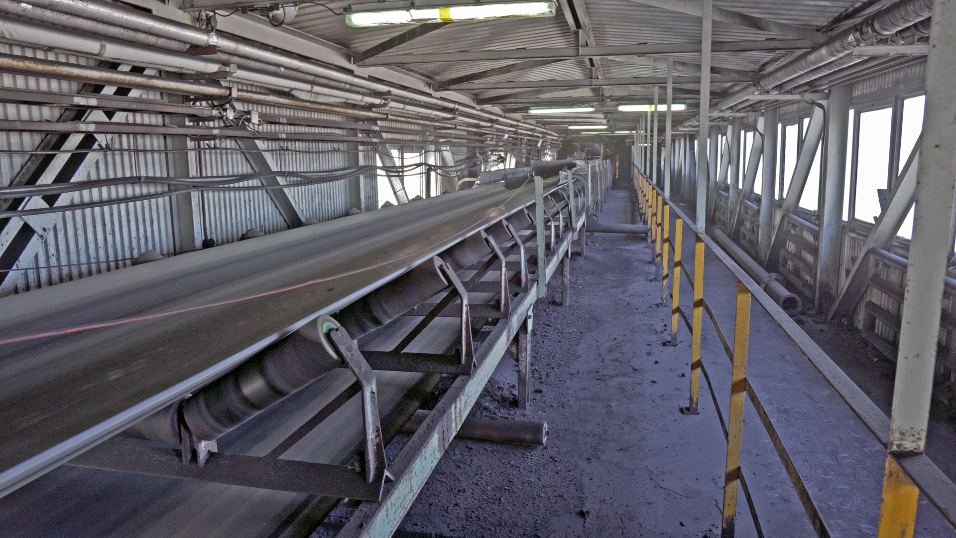 Na zdjęciu widoczny jest przenośnik taśmowy. Przenośnik jest konstrukcją modułową zbudowaną ze stalowych elementów. Na rolkach o kształcie walca osadzona jest taśma. Taśma jest pusta. Przenośnik taśmowy znajduje się w zabudowanym budynku. Po obu stronach łącznika widoczne są okna.