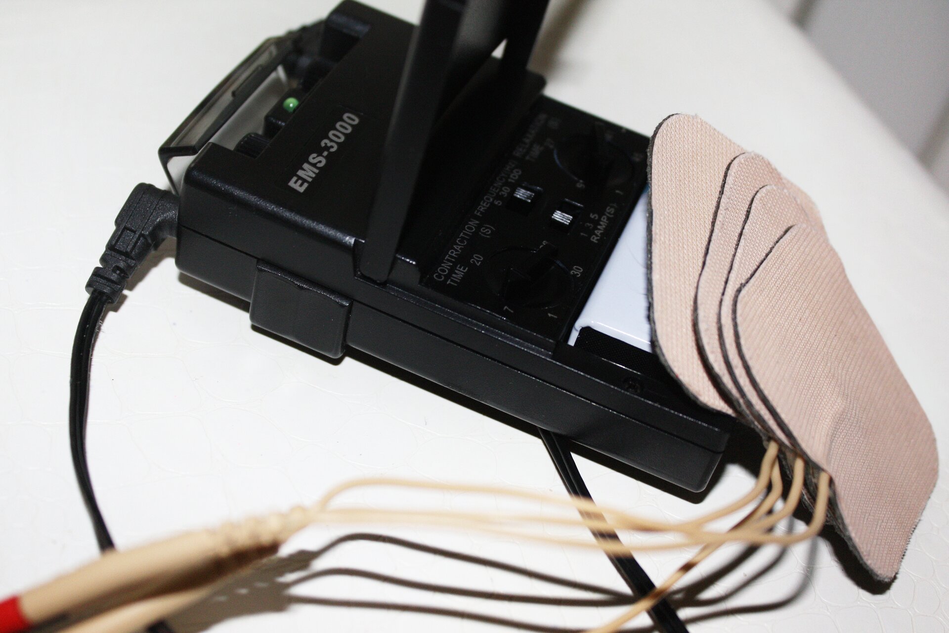 Zdjęcie przedstawia urządzenie do elektrostymulacji. Urządzenie składa się z korpusu w obudowie z czarnego tworzywa sztucznego i czterech elektrod podłączonych do urządzenia za pomocą przewodów. Elektrody są kwadratowe.