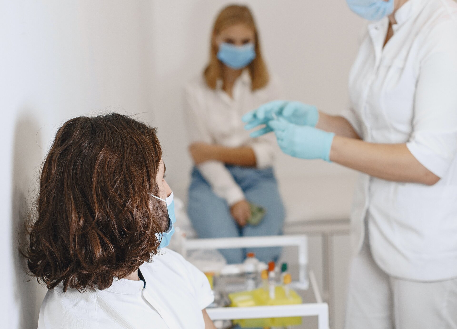 Zdjęcie przedstawia gabinet zabiegowy, w którym znajdują się trzy osoby. Na pierwszym planie widoczna jest siedząca kobieta, w ujęciu od klatki piersiowej w górę, która ma głowę zwróconą w prawo i maseczkę ochronną na twarzy. Na prawo od niej widoczna jest stojąca postać, zwrócona w lewo, ubrana w biały strój pielęgniarski, niebieskie rękawiczki jednorazowe i maseczkę ochronną na twarzy. Osoba przygotowuje zastrzyk dla kobiety siedzącej po lewej stronie. Przed nią widoczny jest wózek z lekami. W tle widoczna jest rozmyta postać kobiety, siedzącej na kozetce.