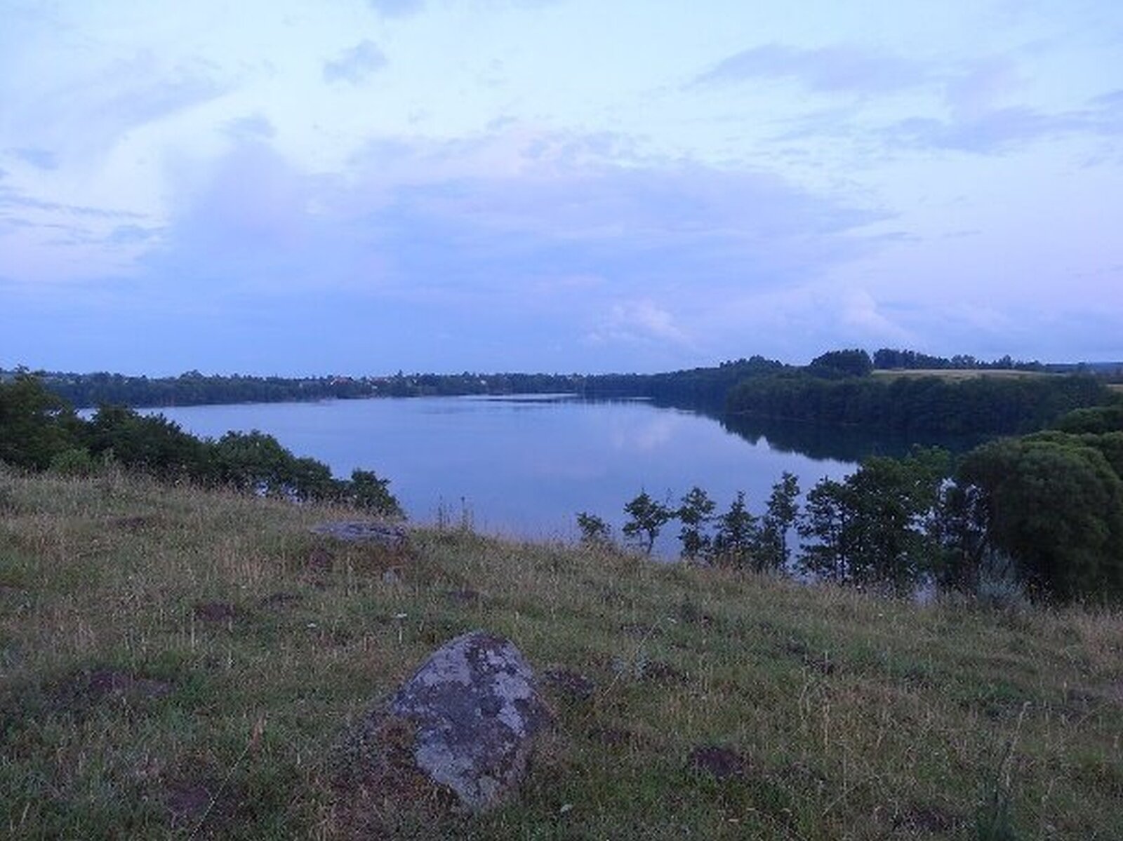 Pośrodku fotografii znajduje się jezioro. Po bokach teren pokryty trawą, w niższych partiach drzewami. Dość strome stoki o wysokości kilku metrów.