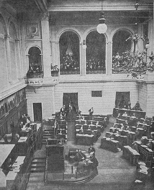 Zdjęcie przedstawia wysoką salę obrad z półkoliście ustawionymi ławami, przy których siedzą ludzie. Nad salą wisi duży żyrandol.