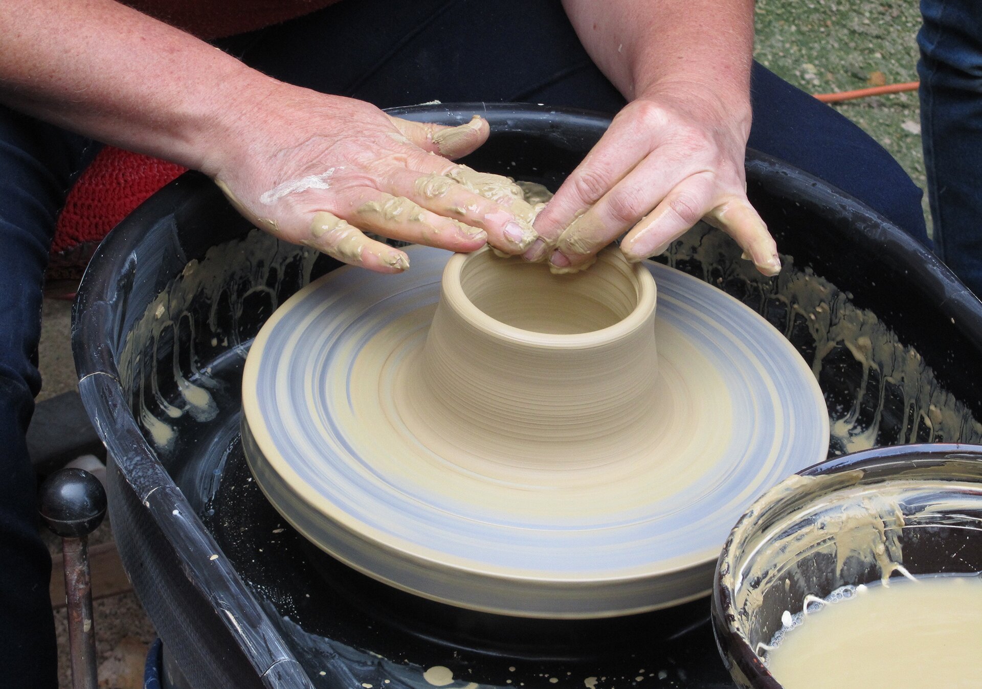 Fotografia przedstawia dwie dłonie ubrudzone gliną, które formują kształt naczynia obracającego się na kole garncarskim. Wokół koła widoczne są zabrudzenia od gliny.