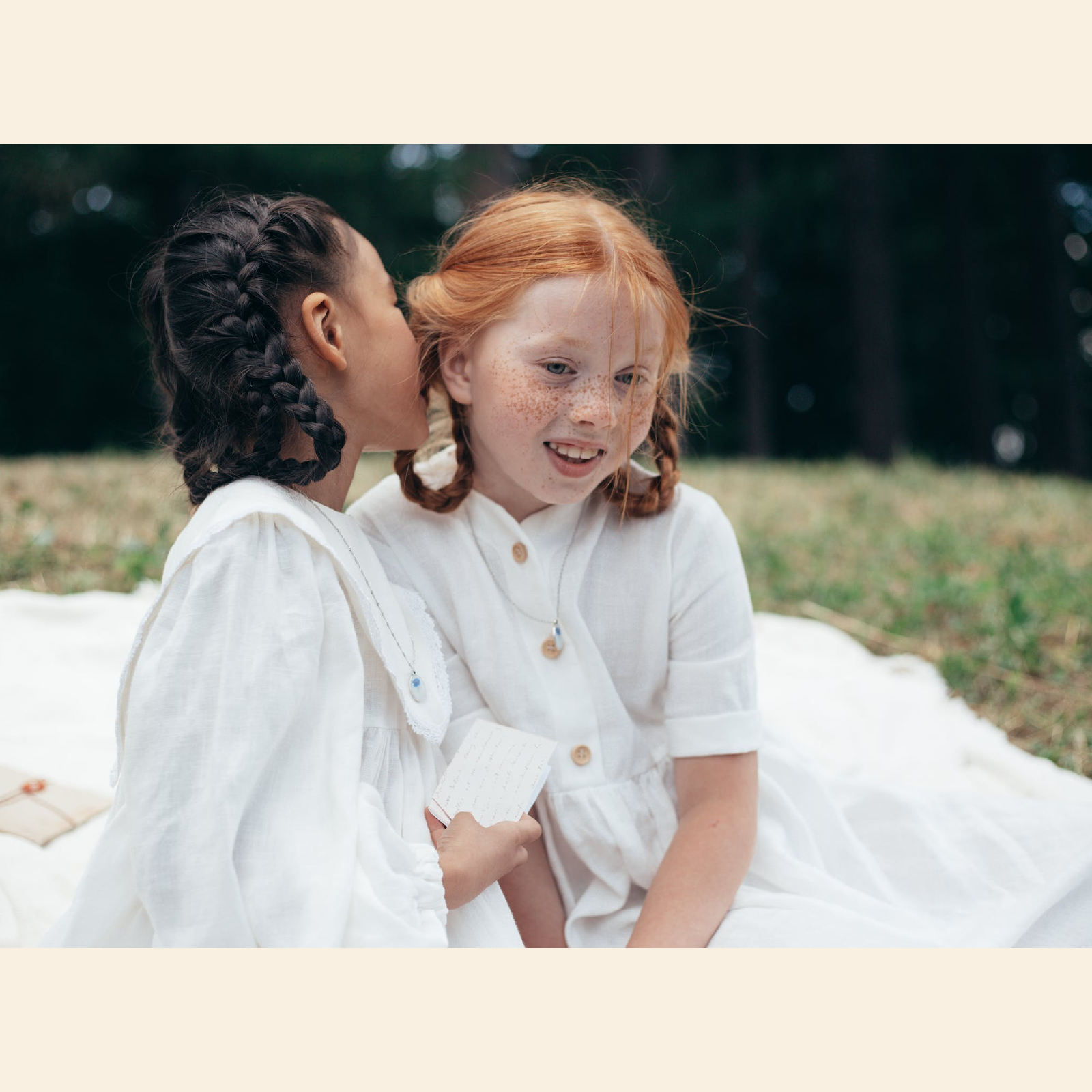 Ilustracja przedstawia dziewczynki w wieku wczesnoszkolnym z zaplecionymi warkoczami. Dziewczynki ubrane są w białe sukienki. Dziewczynka w czarnych włosach szepcze coś na ucho dziewczynce w rudych włosach z piegami.
