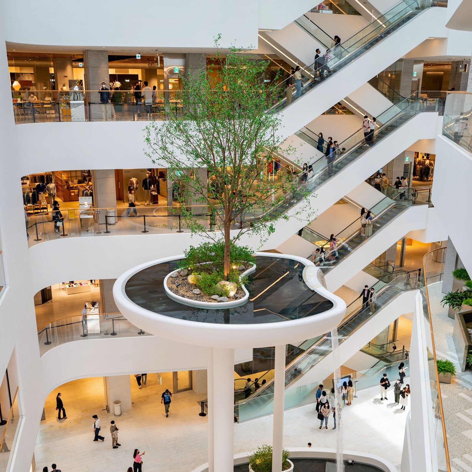 Zdjęcie przedstawia nowoczesną, wielopoziomową galerię handlową. Widoczne są cztery poziomy galerii, które są połączone parami ruchomych schodów jadących w górę i w dół. Na ruchomych schodach widoczni są ludzi jadący w górę i w dół. Na każdym piętrze widoczne są oświetlone witryny sklepowe oraz przechadzający się ludzie. W centralnej części, wokół której rozlokowane są piętra, widoczna jest fontanna w kształcie wyspy, która stoi na dwóch wysokich słupach. Pośrodku wyspy widoczna jest nerkowata powierzchnia wypełniona ziemią i kamieniami, w której rośnie drzewo oraz niskie rośliny. Wokół widoczna jest woda fontanny. Po jej prawej stronie widoczny jest opadający strumień wody. 