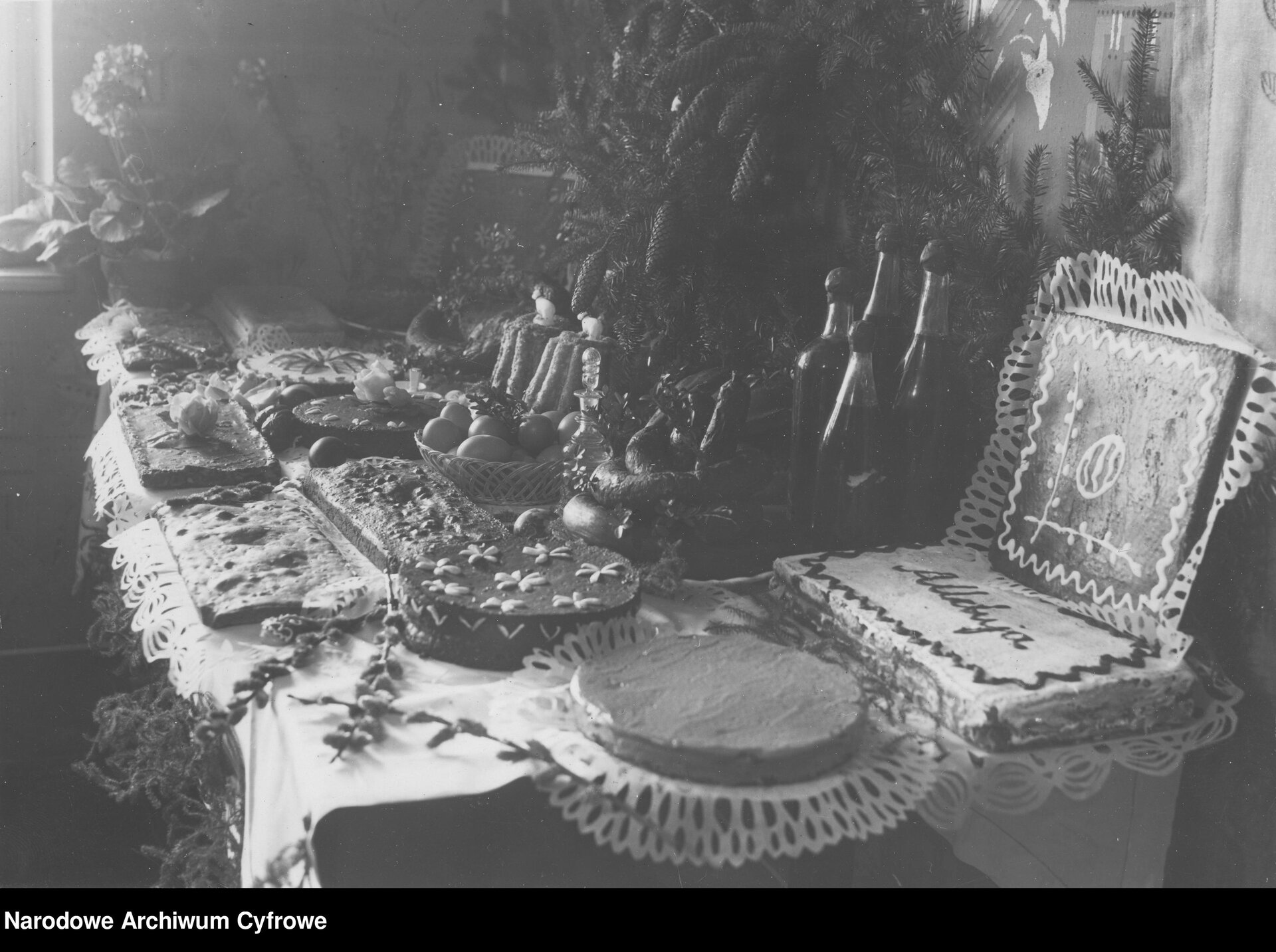 Zdjęcie przedstawia udekorowany wielkanocny stół. Na pierwszym planie widoczne ciasta mazurki okrągłe i prostokątne, dalej placki i babki. Jeden z mazurków ma napis “Alleluja”, inny kwiatki ułożone z migdałów, jeszcze inny narysowaną lukrem pisankę i bazie. Ciasta leżą na białych, wycinankowych serwetkach. Na dalszym planie potrawy wielkanocne, kosz z jajkami, butelki z nalewkami. Stół przystrojony jest iglastym drzewkiem oraz kwiatami w doniczce, z lewej strony fotografii przez okno przebijają się promienie światła słonecznego.