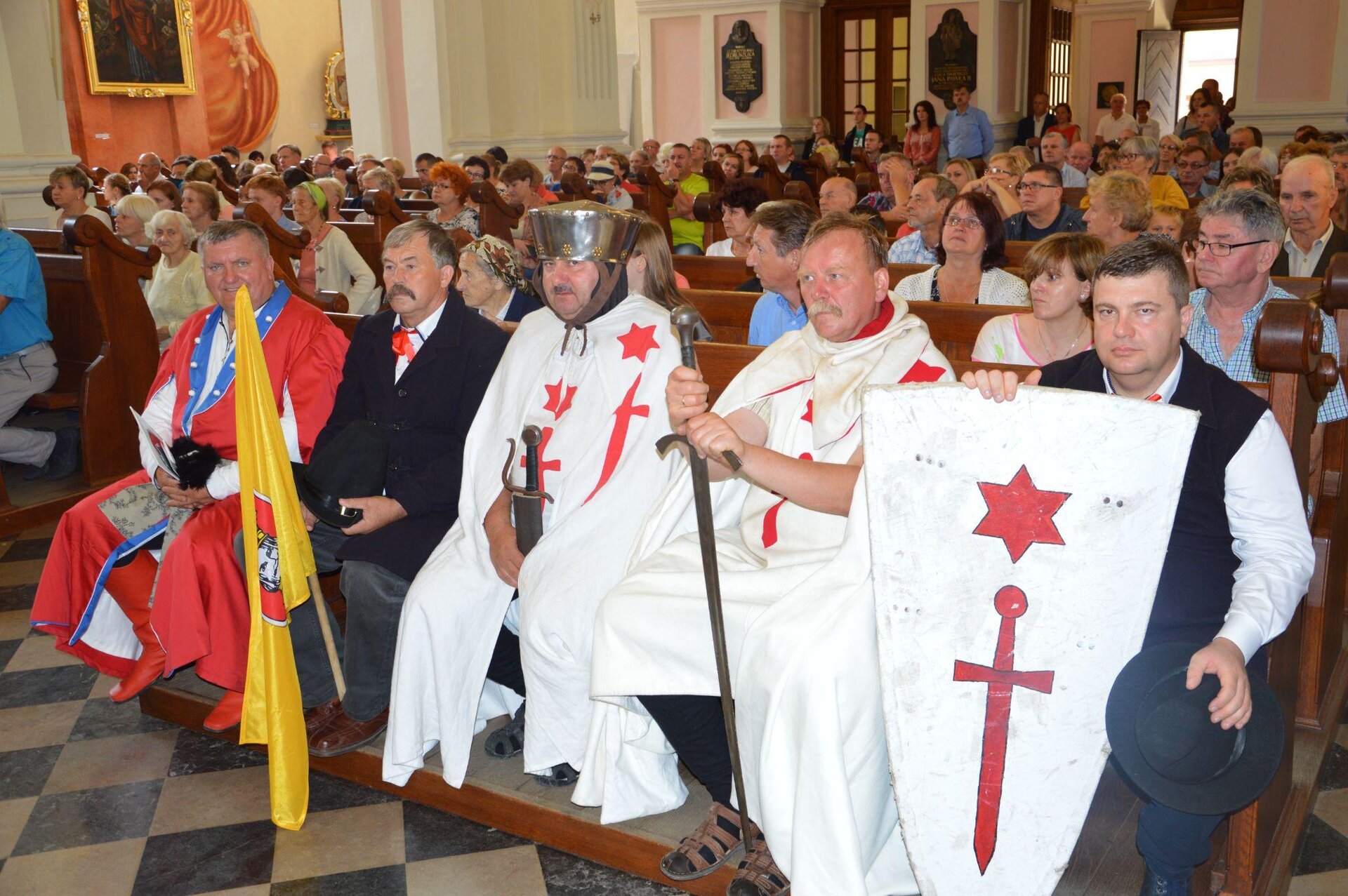 Kliknij, aby powiększyć. Kolorowe zdjęcie przedstawia widok wnętrza kościoła. W drewnianych ławkach siedzą kobiety i mężczyźni. W pierwszym rzędzie widać pięć osób ubranych w stroje z epoki. Od prawej: mężczyzna w białej koszuli i czarnej kamizelce, który przed sobą trzyma dużą białą tarczę z czerwonym znakiem gwiazdy i miecza, obok dwóch mężczyzn w białych płaszczach rycerskich z mieczami w dłoniach, dalej mężczyzna w białej koszuli i czarnym płaszczu z żółtą flagą, ostatni siedzi mężczyzna w czerwonych wysokich butach i czerwonym kontuszu obszytym niebieską lamówką.