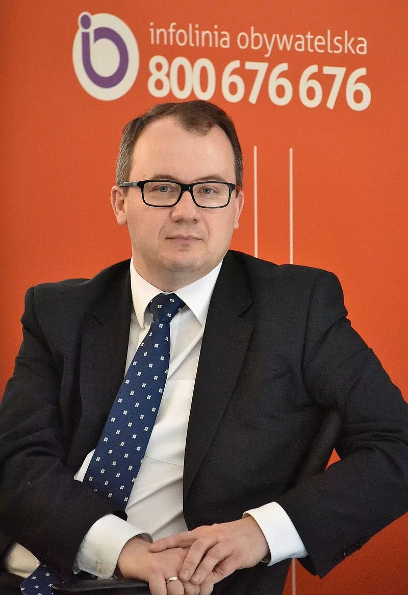Zdjęcie przedstawia mężczyznę w średnim wieku. Twarz mężczyzny jest owalna, włosy ciemne, krótko ostrzyżone. Mężczyzna ma okulary w czarnych, plastikowych oprawkach. Ubrany jest w garnitur, koszulę i krawat. Mężczyzna siedzi na tle ściany z napisem: infolinia obywatelska 800676676.
