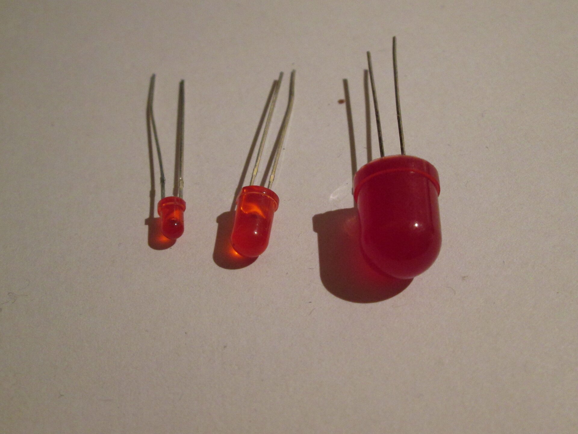 Rys. 5. Zdjęcie przedstawia trzy diody LED w postaci walców w czerwonym kolorze zaokrąglonych w dolnej części. W kolorowych elementach diody znajduje się materiał półprzewodnikowy, po podłączeniu którego do prądu dioda zaczyna świecić. Do diod w górnej części nie zaokrąglonej przymocowane są po dwa druciki, którymi doprowadzany jest prąd elektryczny. Diody leżą jedna obok drugiej. Dioda po lewej jest najmniejsza, a po prawej największa.