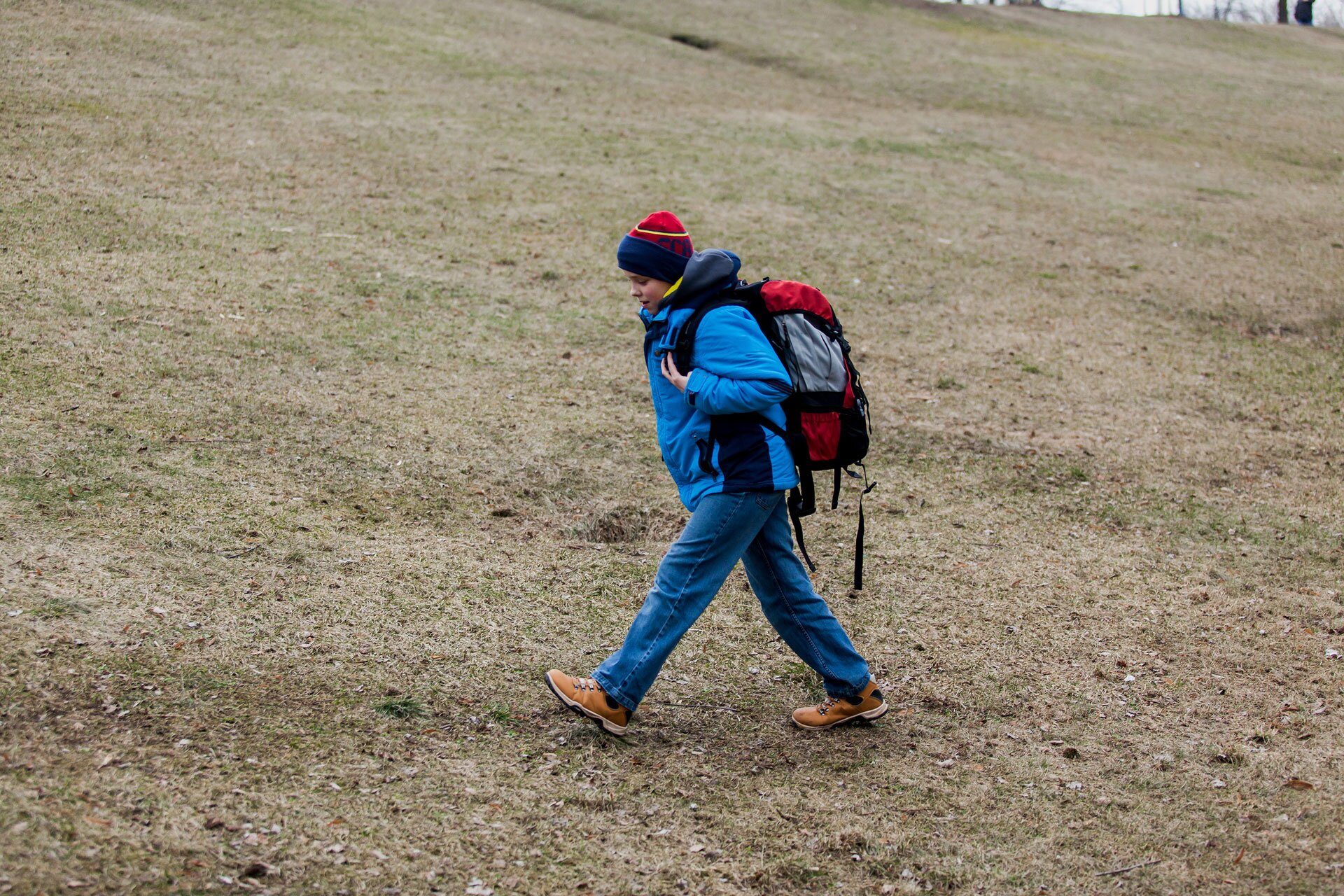 Ilustracja przedstawia idącego pod górę chłopca. Chłopiec idzie po terenie porośniętym niską trawą. Trawa jest brązowa. Chłopiec ma ubraną niebiesko-czerwoną czapkę, niebieską kurtkę i dżinsy, brązowe buty. Na plecach czerwono-szary plecak.