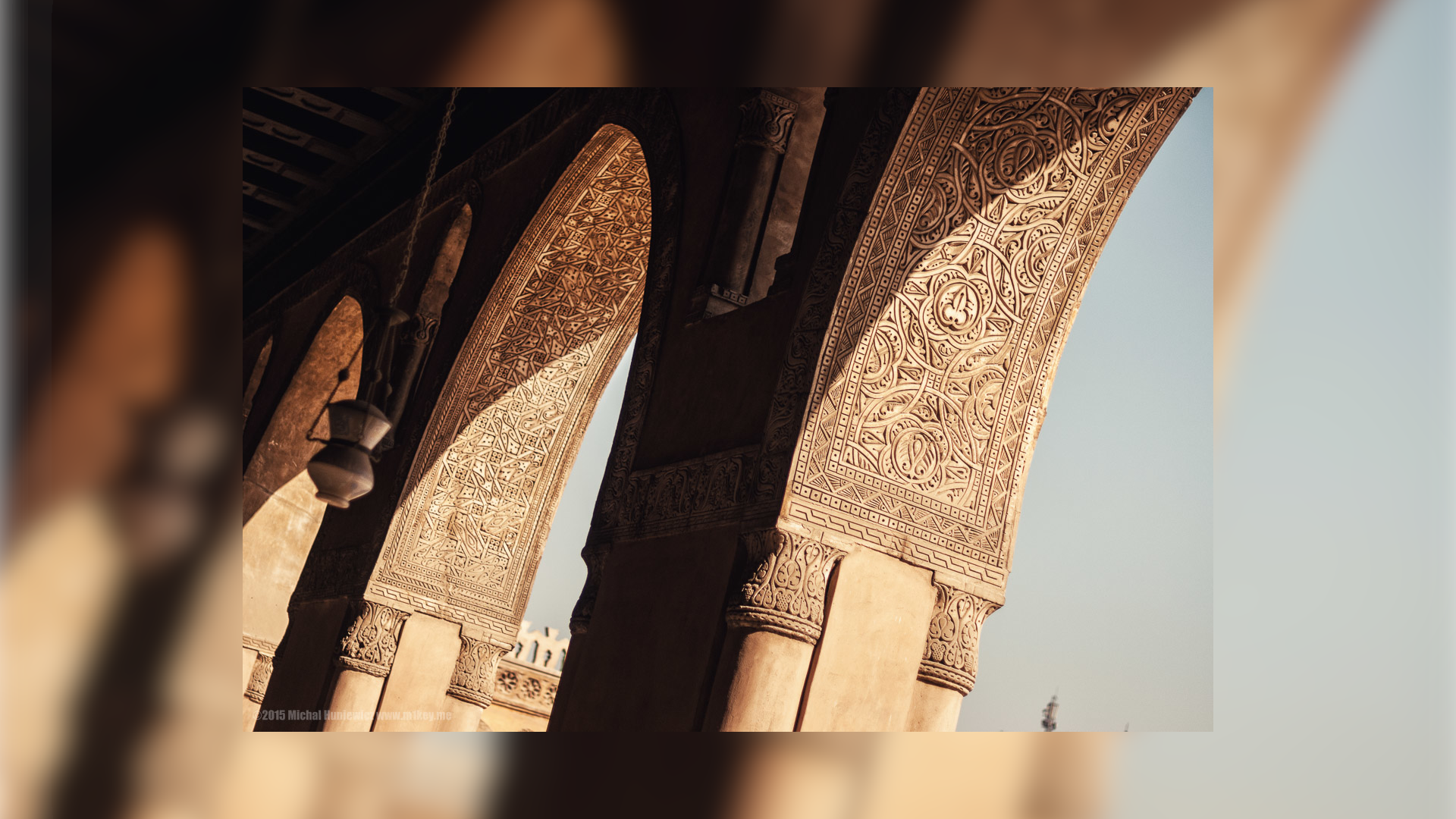 Na zdjęciu znajdują się łuki stanowiące część meczetu. Są one ozdobione licznymi dokładnymi wzorami. Z sufitu wnęki zwisa kadzidło. 