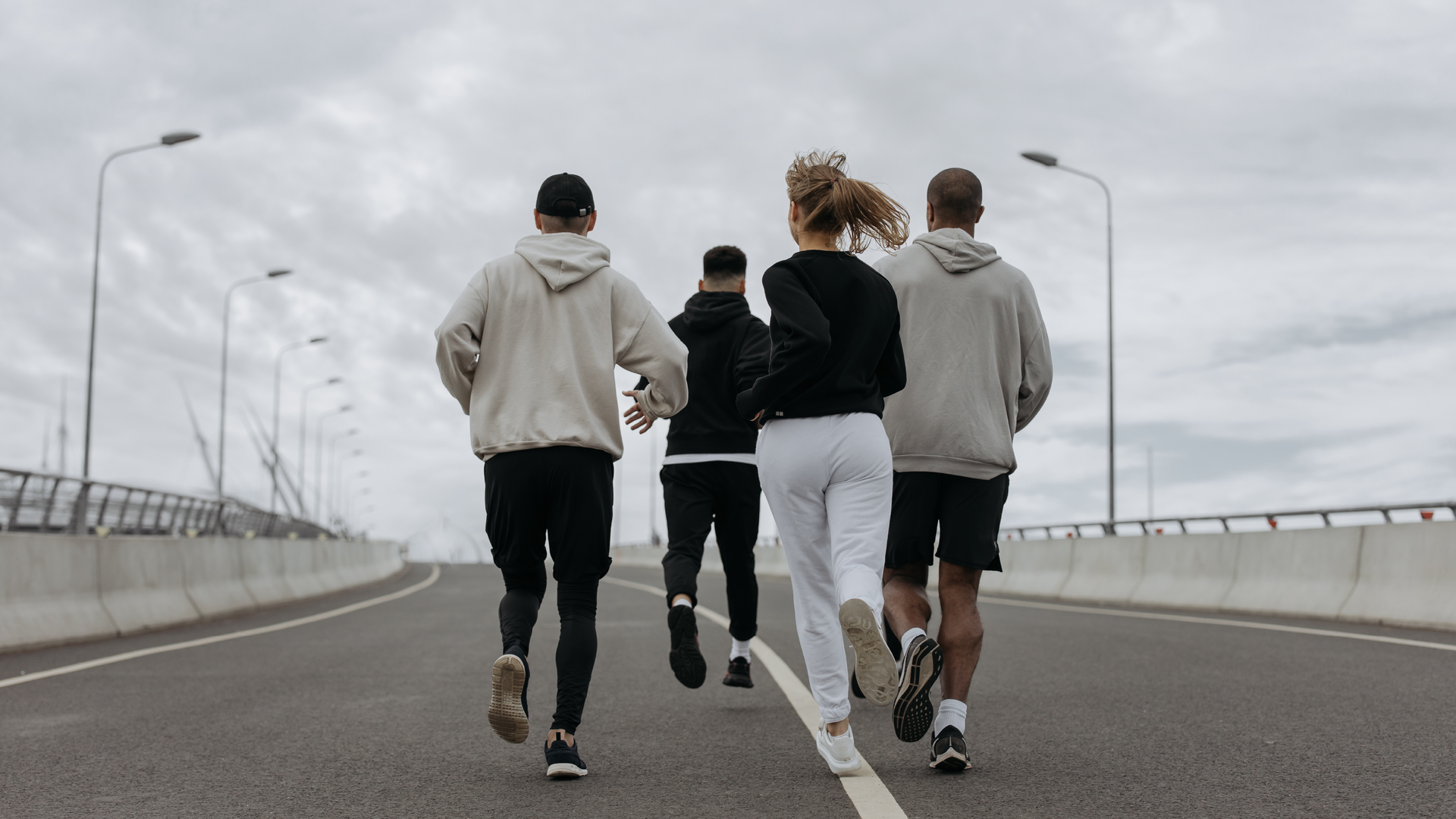 Zdjęcie przedstawia cztery biegnące osoby, dziewczynę i trzech chłopaków. Ubrani są w bluzy, spodnie dresowe, a jeden z chłopaków w krótkie spodenki. Biegacze znajdują się tyłem do kadru, niewidoczne są ich twarze. Biegną szeroką, asfaltową drogą z wymalowanymi ciągłymi liniami po prawej i po lewej stronie oraz przez środek. Po bokach drogi znajdują się betonowe bariery ochronne oraz latarnie.