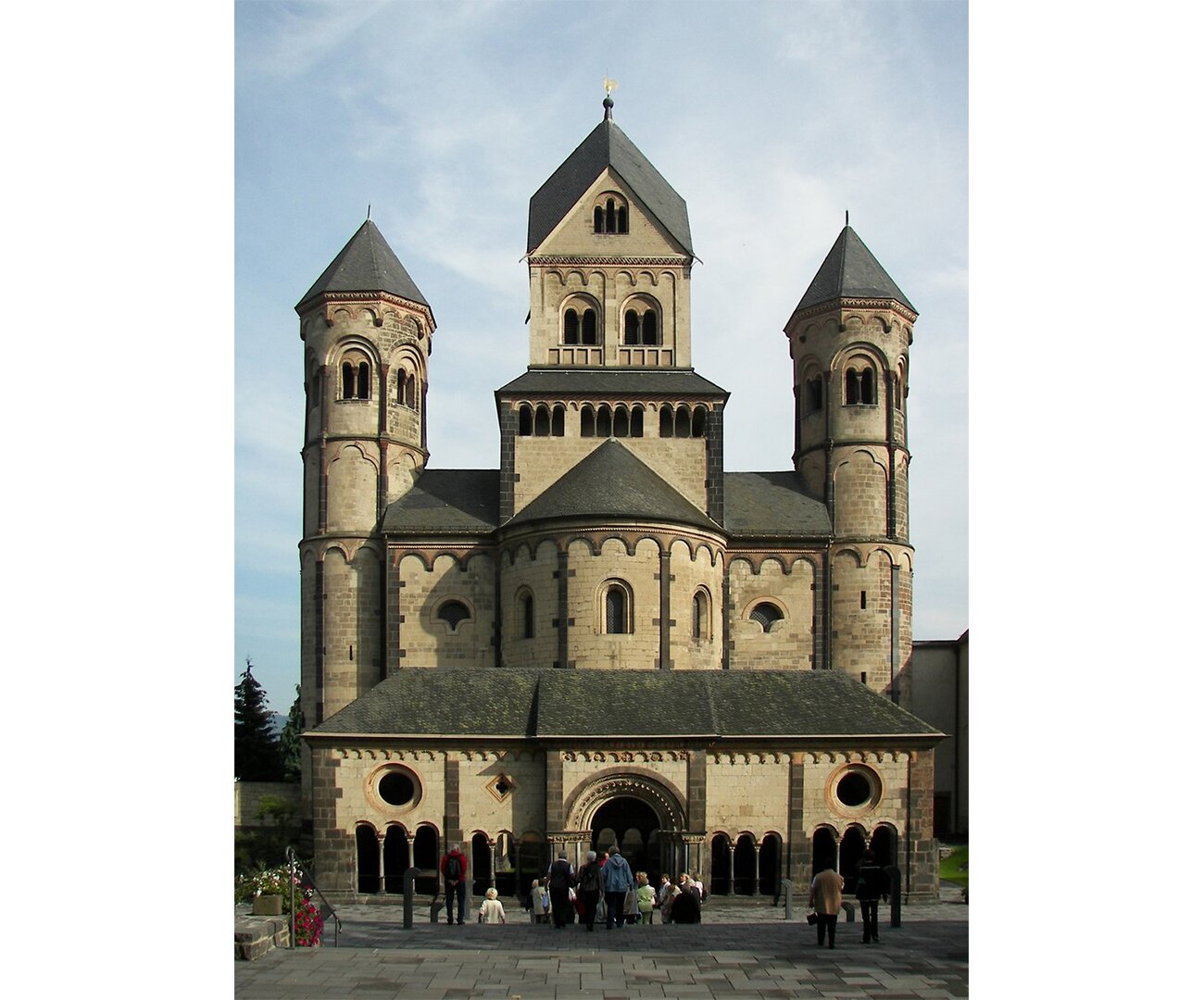 Fotografia barwna przedstawiająca Kościół opactwa Maria Laach, XII wiek (portyk przed kościołem - I ćw. XIII w.) Kościół ma charakterystyczne wyżłobione elementy przy oknach. Na dole u podstawy okna są mniejsze, okrągłe. Budynek ma trzy wieże z czego środkowa jest najwyższa.