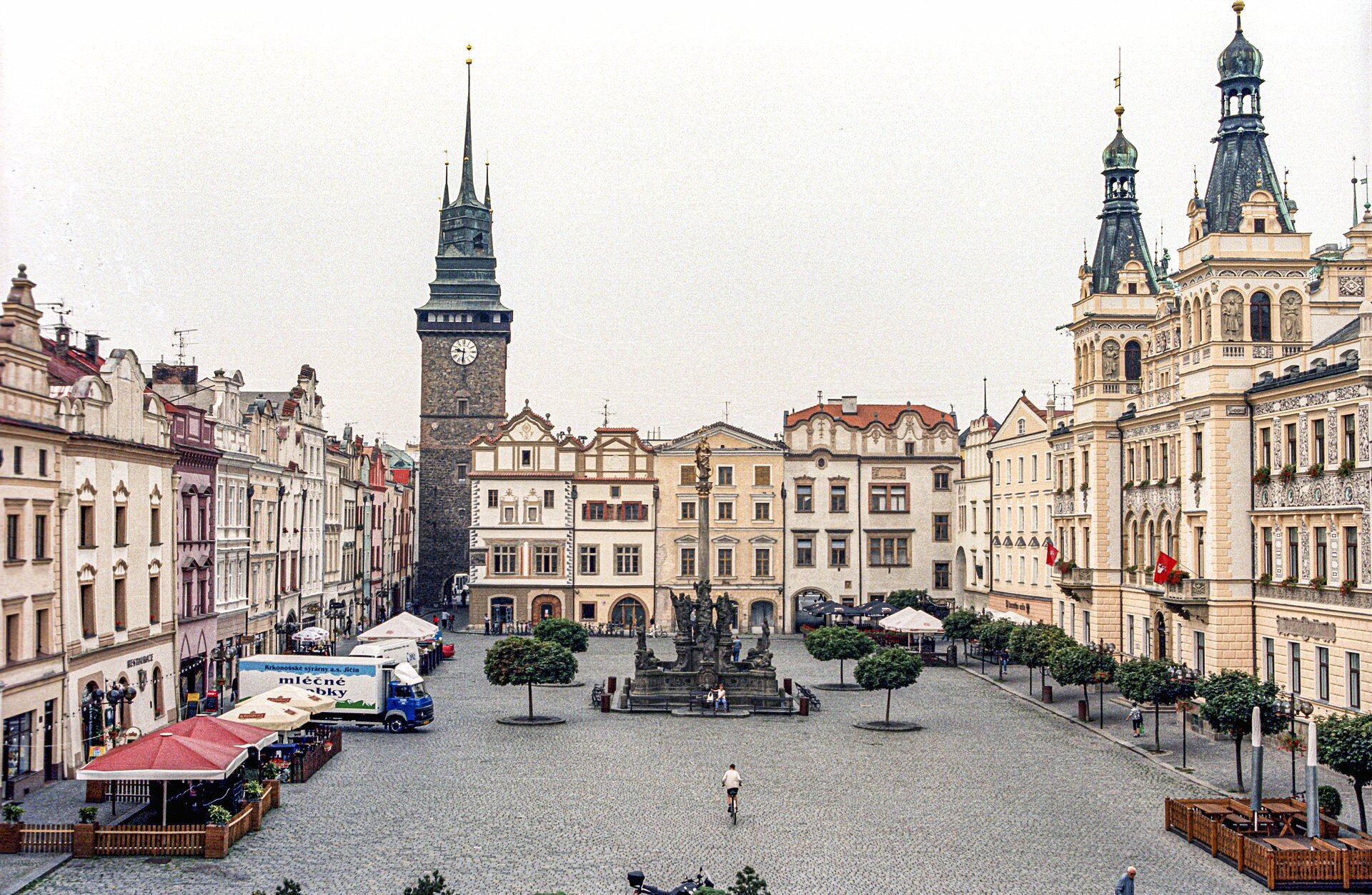 Rynek miejski w Pardubicach - zdjęcie przedstawiające kamienice w jasnym kolorze oraz wieżę koloru ciemnobrązowego z zieloną wieżą. Na środku rynku znajduje się pomnik. 