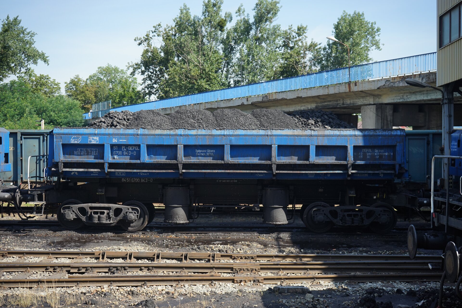 Na zdjęciu widoczny jest załadowany na wagonach węgiel. Węgiel nasypany jest w formie kilku stożków. Spodnia część pryzmy jest szersza i zwęża się ku górze. Za niebieski wagonem widać drzewa.Poniżej zdjęcia widoczny jest przycisk multimedialny z nagraniem audio. Treść nagrania jest zgodna z tytułem zdjęcia.
