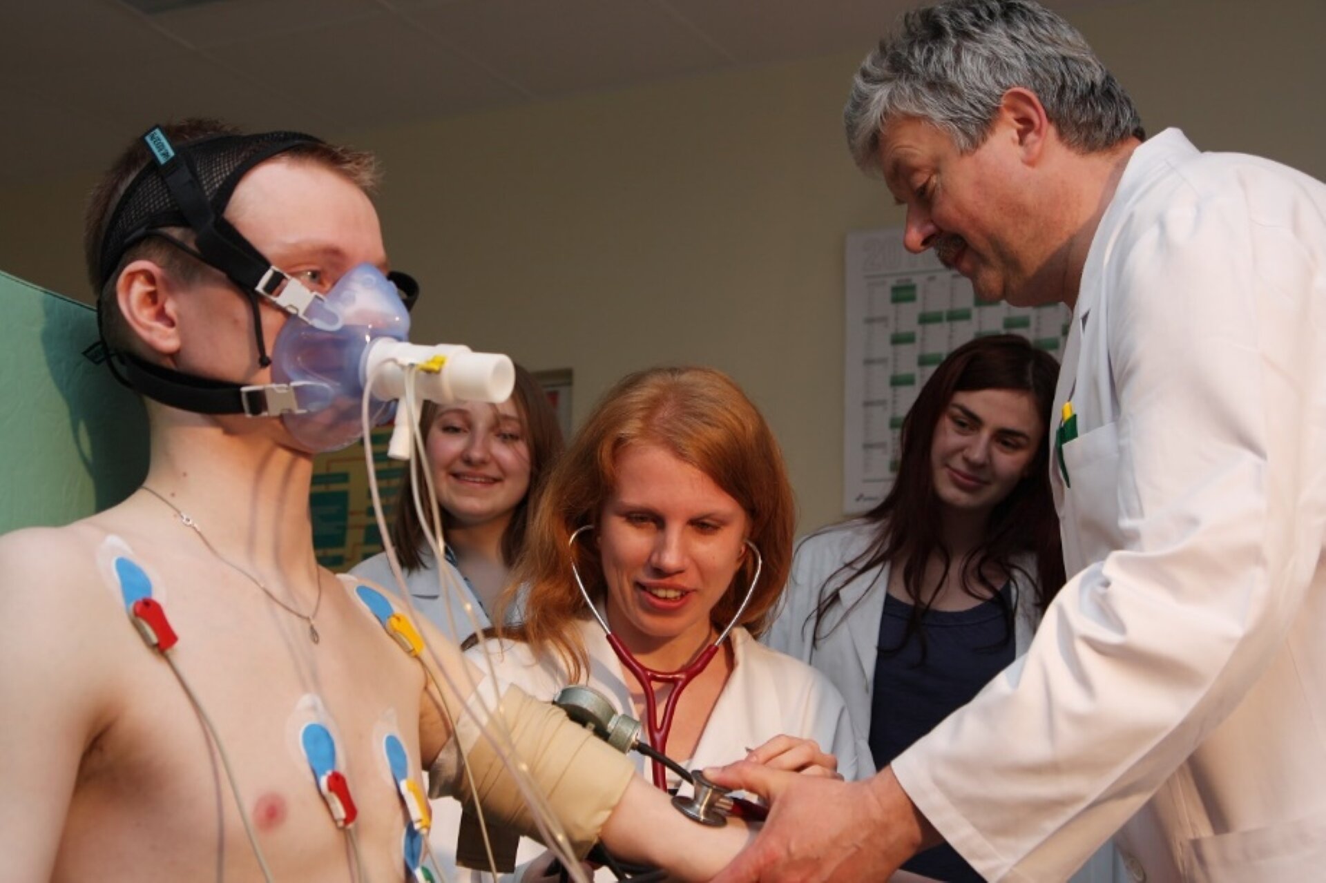 Rys. a. Ilustracja przedstawia zdjęcie, na którym widoczny jest pacjent w gabinecie lekarskim. Młody mężczyzna siedzący na fotelu medycznym poddawany jest badaniu. Do gołej skóry na klatce piersiowej ma przyklejone czujniki połączone białymi przewodami z innym urządzeniem sczytującym wyniki pomiarów. W ten sposób najprawdopodobniej mierzone są parametry oddechowe. Na lewym ręku widoczna jest jasna opaska uciskowa. Stojący naprzeciwko mężczyzny lekarz za pomocą ciśnieniomierza dokonuje pomiaru ciśnienia krwi pacjenta, na jego lewym ręku poniżej opaski.  Lekarz ubrany jest w biały fartuch. Pacjent na twarzy ma plastikową maskę z rurką podłączonymi białymi rurkami. Służy ona do pomiaru pojemności płuc lub składu wydychanego powietrza. W tle widać trzy młode kobiety ubrane w białe fartuchy lekarskie. Kobiety przyglądają się badaniu w ten sposób ucząc się poprawnego wykonywania pomiarów.