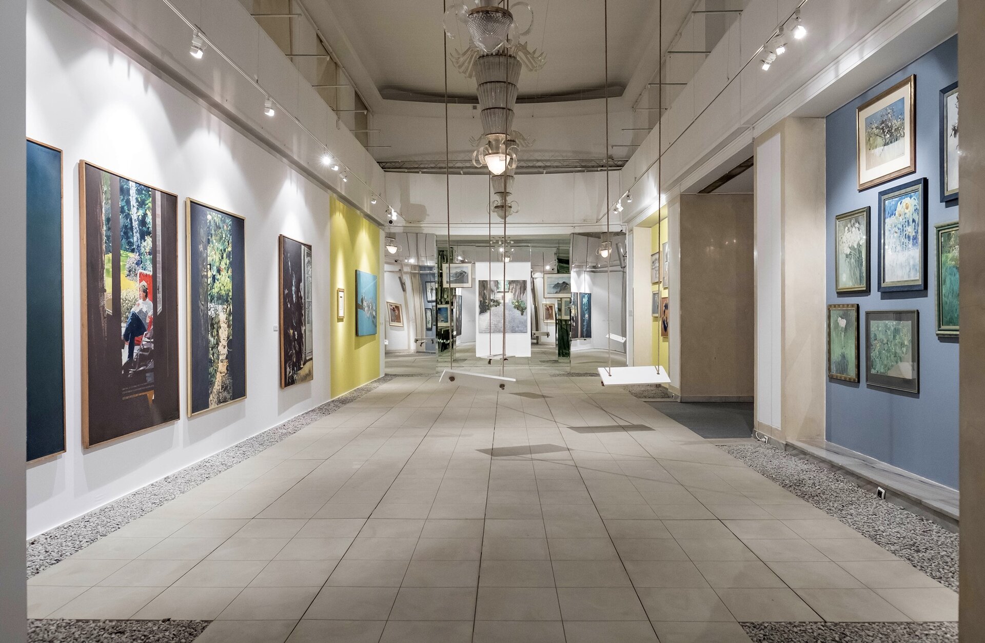 Na zdjęciu znajduje się korytarz z szarą podłogą. Ściany mają różne kolory: biały, żółty, beżowy, fioletowy. Wiszą na nich obrazy o zróżnicowanej wielkości. Nad dziełami znajdują się małe, białe lampy, a na środku sufitu są duże, ozdobne żyrandole. Dookoła żyrandoli powieszone są na długich linach 3 białe huśtawki. W tle znajdują się kolejne obrazy. 