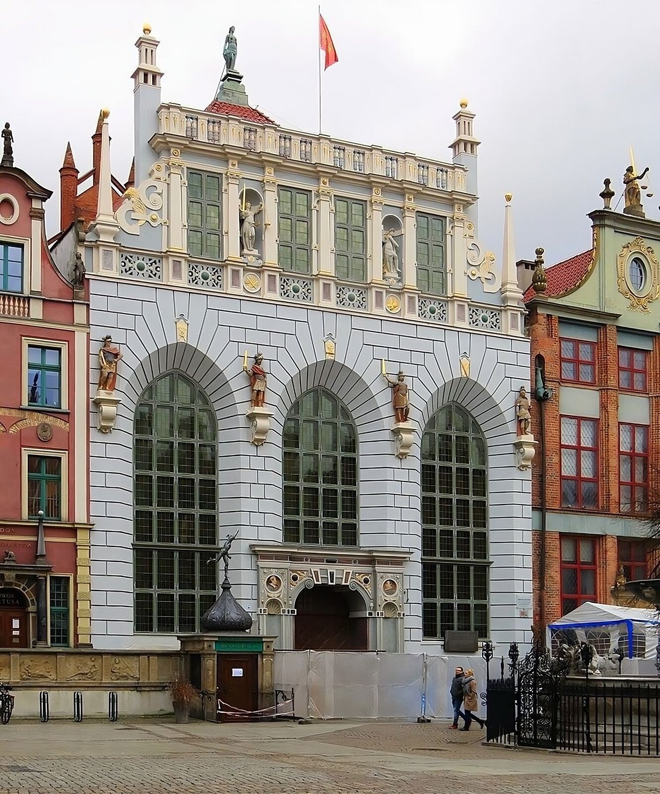 Zdjęcie przedstawia widok współczesny na fasadę Dworu Artusa w Gdańsku. Konstrukcja renesansowa, z okazałym portalem i medalionami królów polskich Zygmunta III Wazy i Władysława IV Wazy. Medaliony ulokowane w górnych płytach narożnych pod fryzem, flankują po obu stronach portal wejściowy. Trzy ostrołukowe okna dominują od części parterowej ku szczytowi. Między nimi cztery figury bohaterów z dziejów mitologii greckiej, rzymskiej i historii antycznej. U szczytu budynku osłaniająca dwuspadowy dach attyka z prostokątnymi lukarnami, przedzielona we wnękach figurami postaci żeńskich - alegorii. Na szczycie dachu figura postaci żeńskiej oraz łopocząca na maszcie nad attyką flaga z herbem Gdańska. Po prawej stronie dworu Artusa Dom Ławy - siedziba dawnego sądu miejskiego, po lewej jedna z kamienic w stylu klasycystycznym. U dołu ilustracji ogrodzenie prowadzonych prac budowlanych i fragment kratownicy osłaniającej basen Fontanny Neptuna.  