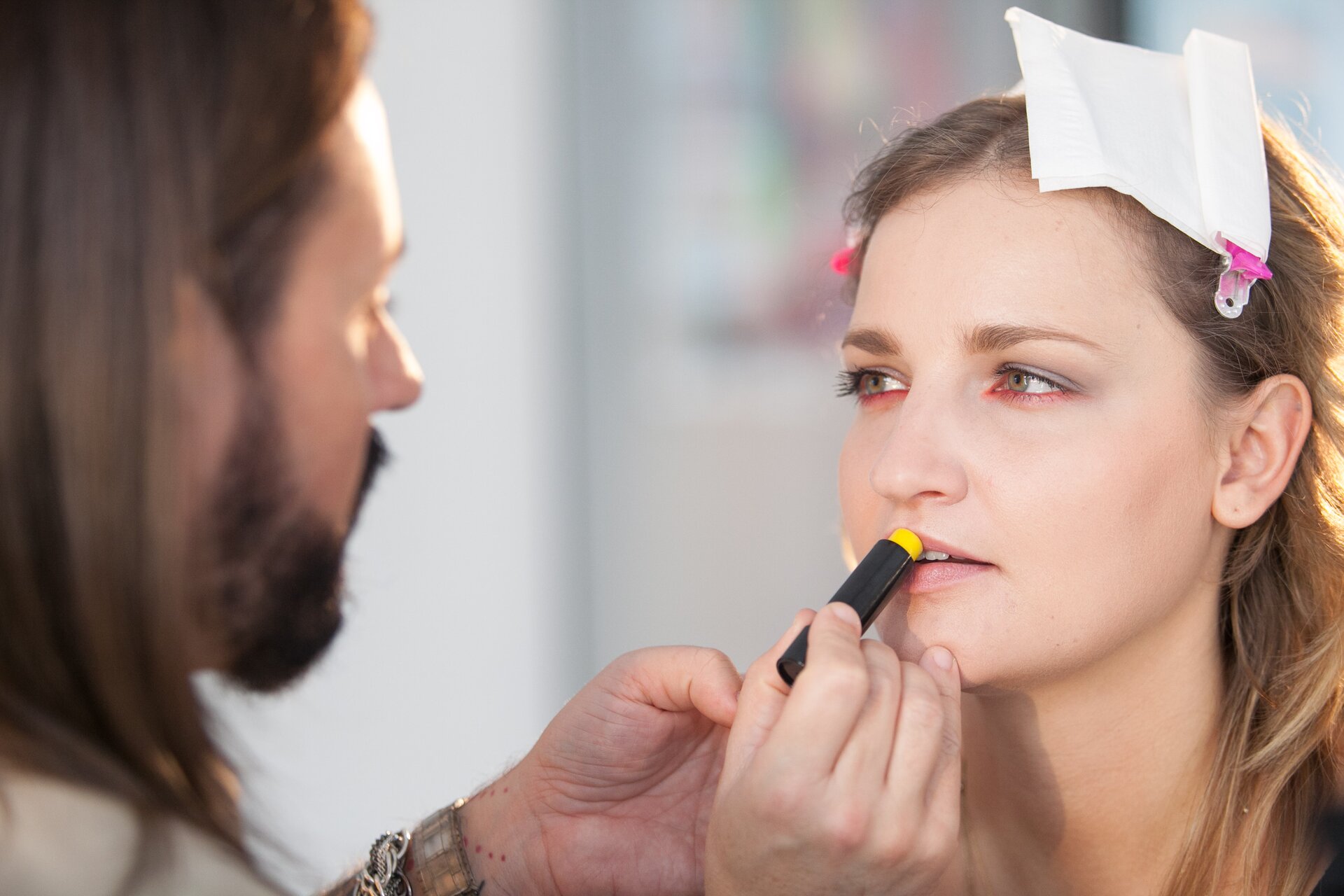 Fotografia prezentuje młodą kobietę o długich, blond włosach, które spięte są przy twarzy za pomocą różowych spinek. Jedna z nich przytrzymuje również białą chusteczkę. Przed kobietą stoi mężczyzna o długich, brązowych włosach oraz brodzie. Mężczyzna wykonuje kobiecie makijaż. Przedstawiony jest w momencie nakładania na jej usta żółtego balsamu w czarnym opakowaniu. Usta kobiety są delikatnie rozchylone, a jej wzrok kieruje się na wizażystę. Tło za ludźmi jest jasne i rozmazane.