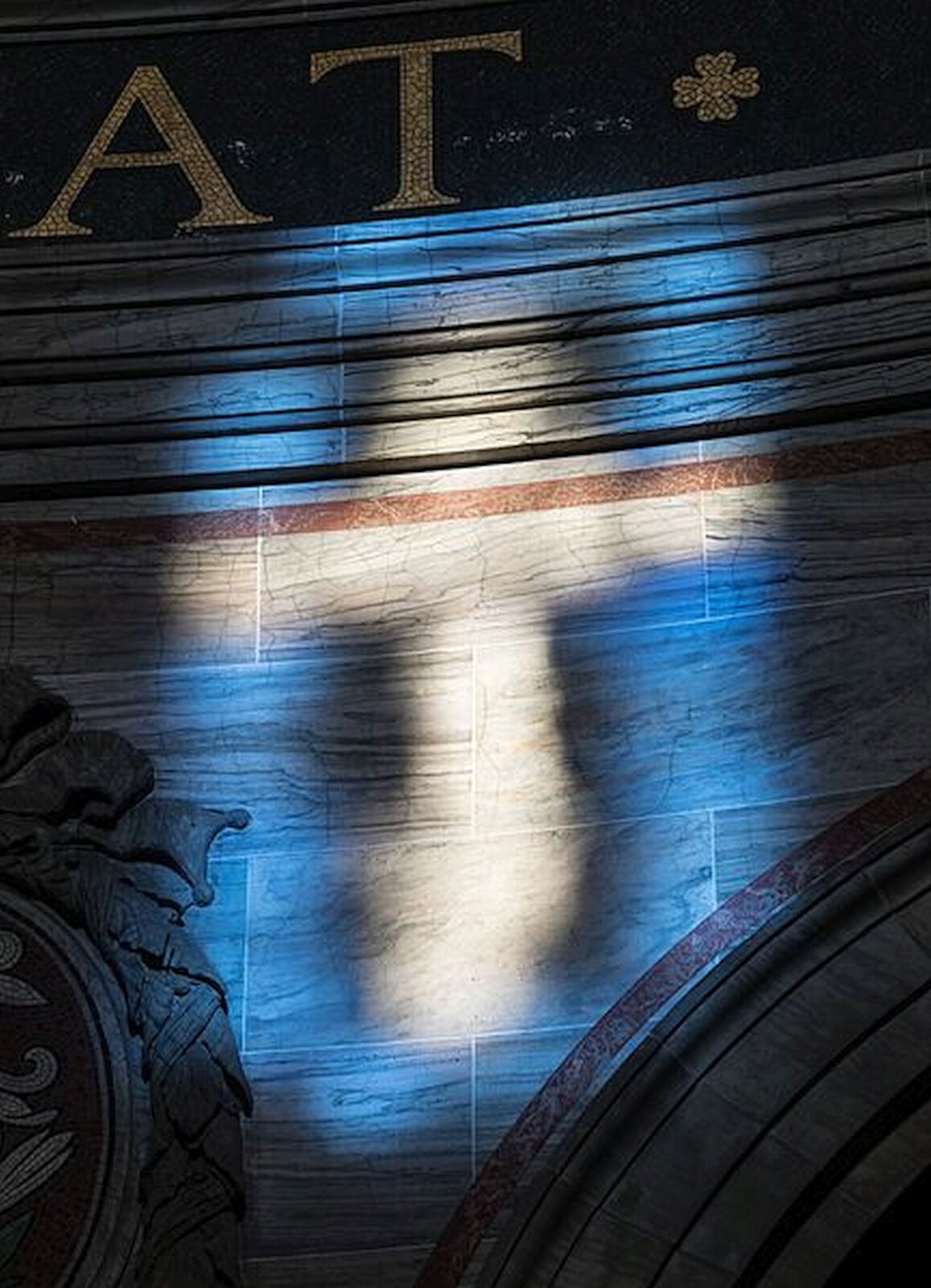 Fotografia przedstawia fragment witraża, przez który przenika światło. Na niebieskim tle ukazany jest rozświetlony, jasny krzyż o rozmazanych konturach.