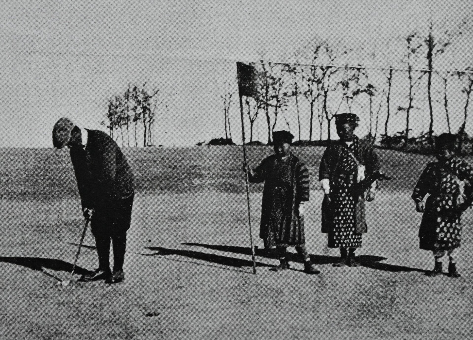 Czarno białe zdjęcie przedstawia mężczyznę stojącego na polu do gry w golfa. Trzyma on w dłoniach kij i dotyka nim piłeczkę. Z boku widoczne są trzy osoby przyglądające się grze. 