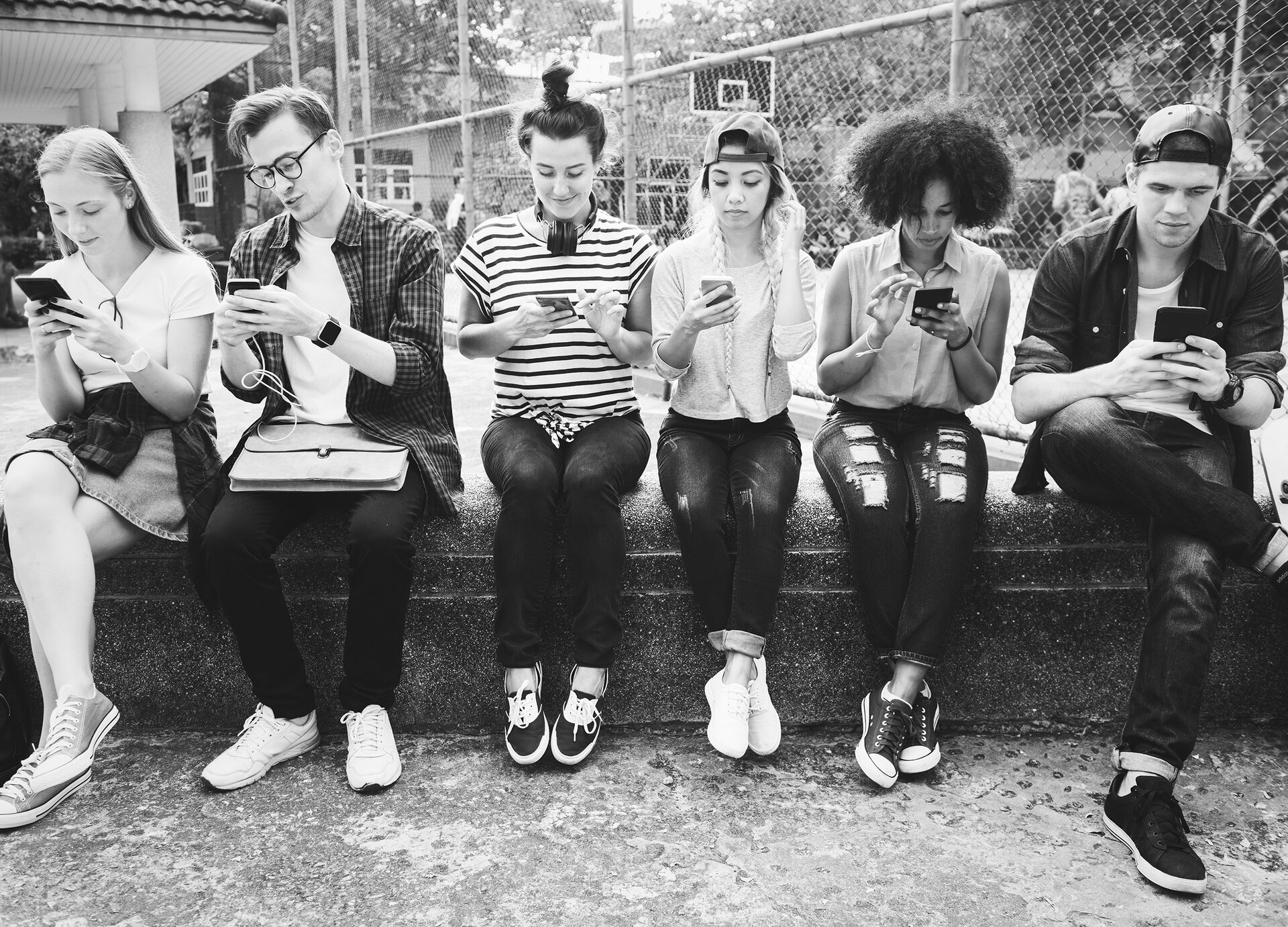  Czarno‑białe zdjęcie przedstawia sześć młodych osób siedzących na murku przed szkołą. Wszyscy ubrani są w niezobowiązujące stroje: dżinsy, bluzki, luźne koszule. Wszyscy mają pochylone głowy i patrzą na telefony komórkowe, które trzymają w rękach. W tle widoczna jest siatka, a za nią boisko, a po lewej stronie widoczny jest fragment zadaszenia nad wejściem do budynku. 
