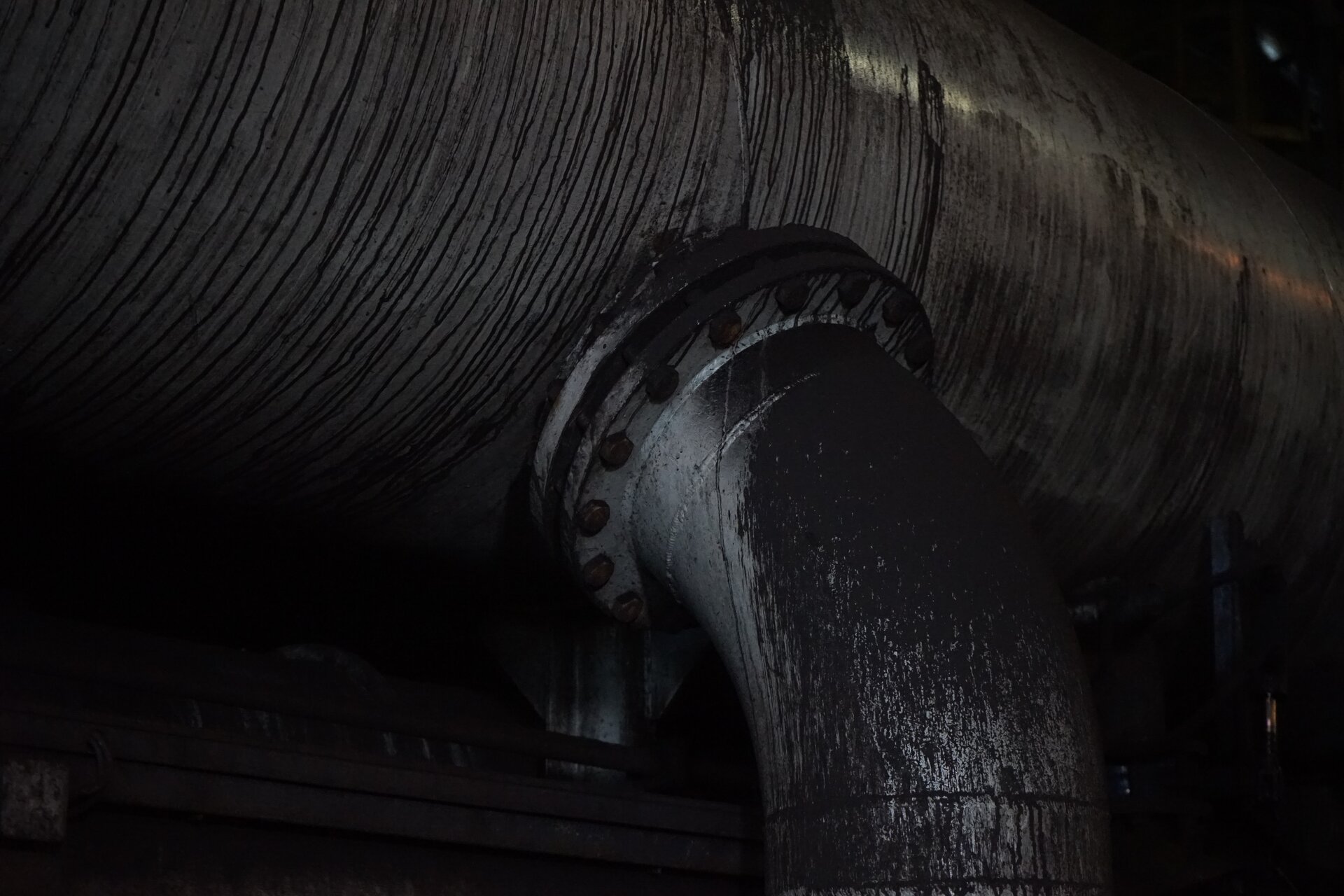 Zdjęcie przedstawia fragment rurociągu doprowadzającego wodę do osadzarki. Jest on poprowadzony od dołu do góry, a następnie wygięty w prawą stronę, gdzie za pomocą łączenia kołnierzowego zamocowany jest do dna skrzyni roboczej osadzarki. Poniżej zdjęcia znajduje się przycisk multimedialny z nagraniem audio. Treść nagrania jest zgodna z tytułem zdjęcia.