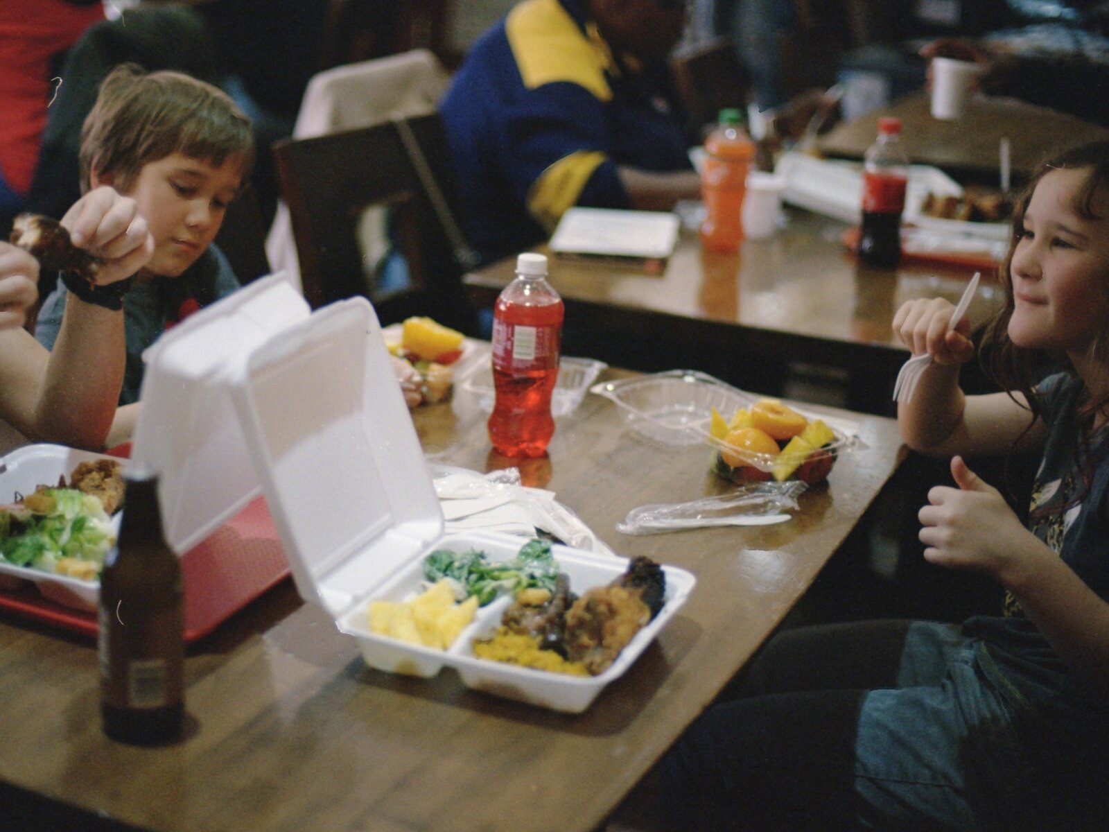 Zdjęcie przedstawia siedzące przy stolikach dzieci, które jedzą lunch. Na prostokątnych, drewnianych stołach widoczne są jednorazowe pudełka, w których znajduje się przygotowane jedzenie, sztućce jednorazowe, butelki z napojami, jednorazowe pojemniki z owocami. 