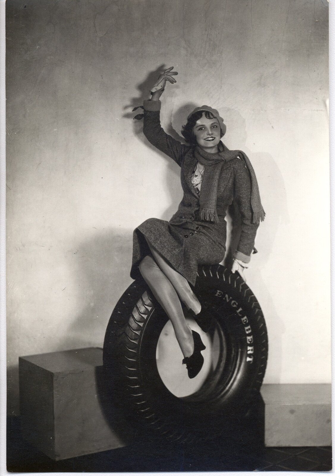 Na zdjęciu reklama opon pochodząca z 1931 roku. Elegancka kobieta w garsonce, z szalem na szyi i beretem na głowie, siedzi uśmiechnięta na oponie. Lewą ręką trzyma się opony, prawą trzyma ku górze - dłoń jest rozwinięta, by pokazać satysfakcję z reklamowanego produktu. 