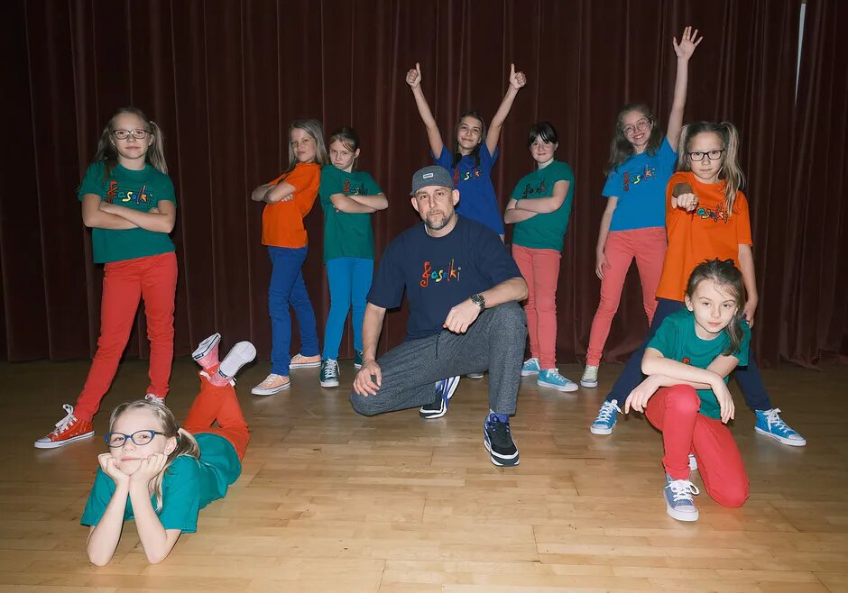 Na zdjęciu, w centrum znajduje się mężczyzna, ubrany w luźne ubrania i czapkę z daszkiem. Wokół niego stoją w różnych pozach dziewczynki, ubrane w kolorowe, podobne spodnie, koszulki i trampki. Za nimi znajduje się kotara.