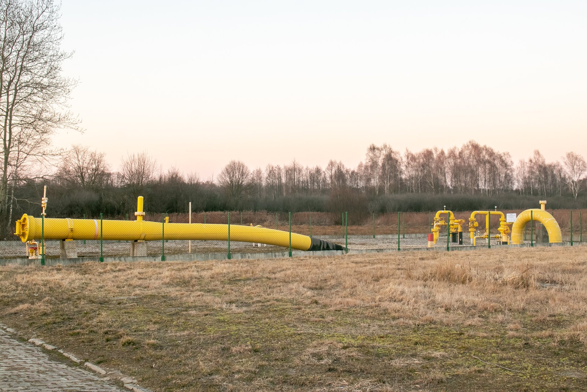 Na zdjęciu widoczny jest rurociąg gazowy wraz z zasuwami odcinającymi na gazociągu przemysłowym. Po lewej stronie zdjęcia widać dużą stalową rurę w kolorze żółtym. Rura jest o przekroju okrągu. Na górze rurociągu widać dwa elementy rurowe które są prostopadłe do niego. Rurociąg posadowiony jest na betonowych prostopadłościennych klockach. Rurociąg zapada w prawą stronę wchodząc stopniowo pod ziemię. Za rurociągiem w niewielkiej odległości widać układ rurociągów z zasuwami odcinającymi na gazociągu przesyłowym. Widoczny na zdjęciu struktura przedstawia układ dwóch rur wychodzących z ziemi które są połączone ze sobą. Układ tworzy kształt obróconej litery U. Rura łączą dwie wychodzące z ziemi rury jest połączona przy użyciu połączenia kołnierzowego. Poniżej połączenia znajdują się zawory z kurkami o kształcie dużych czerwonych okręgów. Po środku rury widoczny jest okrągły wskaźnik ze wskazówką. Na zdjęciu znajdują się dwa takie układy. Układy są połączone przy użyciu elementu z dużym okrągłym kurkiem o kształcie koła. Wszystkie elementy systemu są zabezpieczony ogrodzeniem.