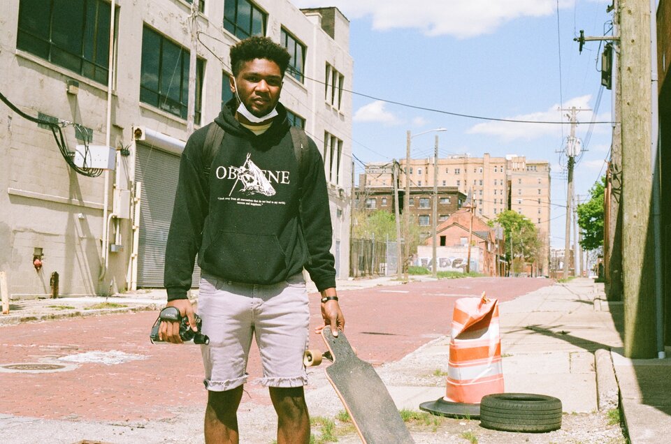 Na zdjęciu znajduje się czarnoskóry młody mężczyzna w czarnej bluzie i białych krótkich spodenkach. Ma kędzierzawe włosy, pod broda ma opuszczoną maseczkę chirurgiczną. Stoi pośrodku ulicy, ręce ma opuszczone. W prawej trzyma małą kamerę, w lewej deskorolkę. Nieco na prawo stoi pogięty drogowy słupek znacznikowy i leży opona. W tle są ściany budynków, wiszą tez kable elektryczne. Dalej w tle dom jednorodzinny, brązowy blok, a jeszcze dalej duży blok w kolorze łososiowym.