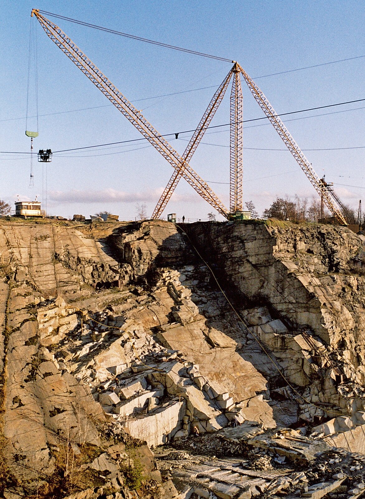 Zdjęcie kamieniołomu. Ma kilkadziesiąt metrów głębokości, jego ściany składają się ze spękanych skał. U góry stoi dużych rozmiarów dźwig.