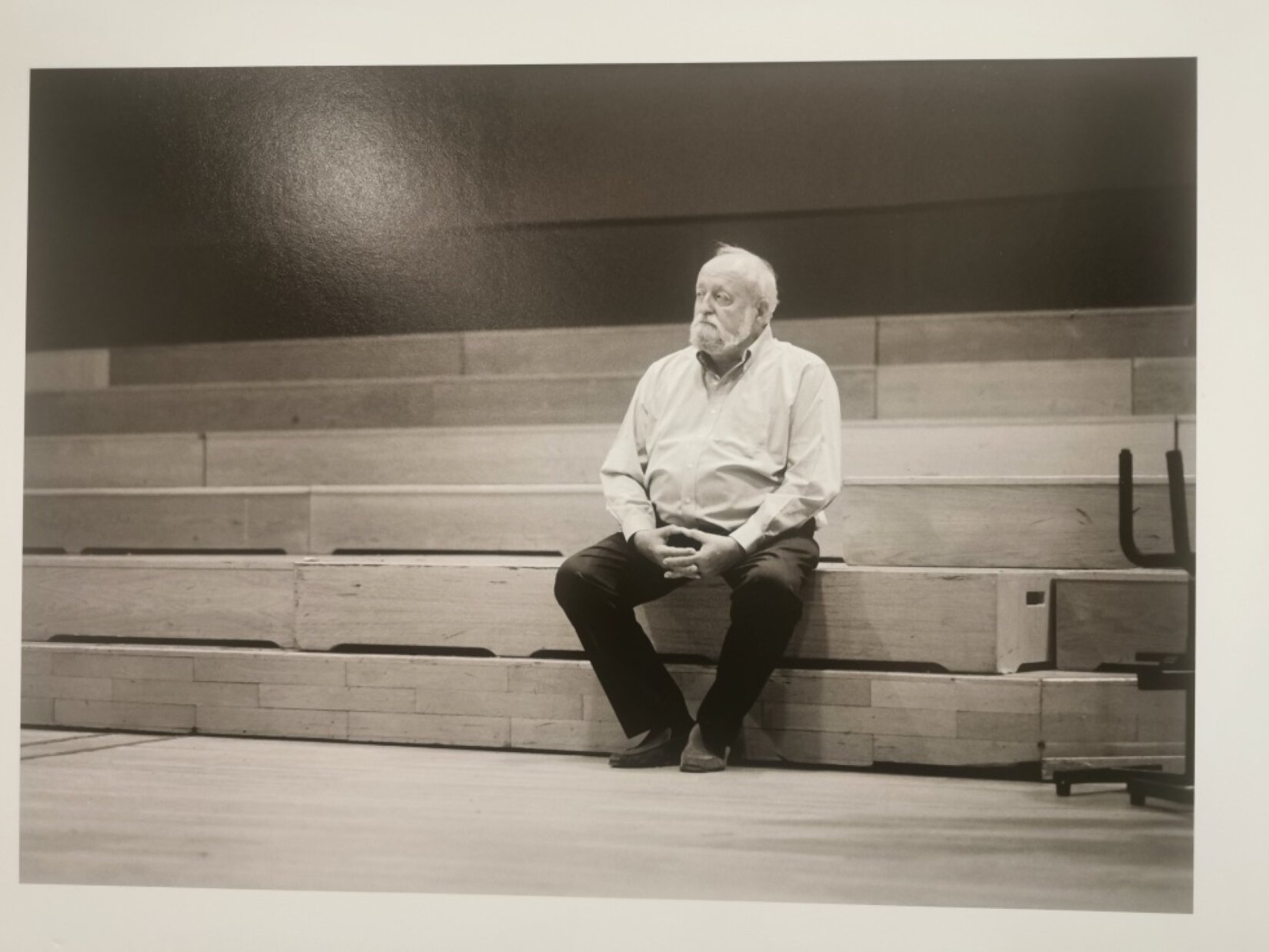 Zdjęcie Krzysztofa Pendereckiego. Na głównym planie znajduje się mężczyzna, który siedzi na szerokich schodach. Ma krótkie włosy, brodę i wąsy. Ubrany jest w koszulę z długim rękawem, długie spodnie i buty. Ręce ma opuszczone, a palce dłoni splecione. Jedynie jego kciuki stykają się opuszkami. W tle znajduje się ściana. Portret jest wykonany w odcieniu sepii.