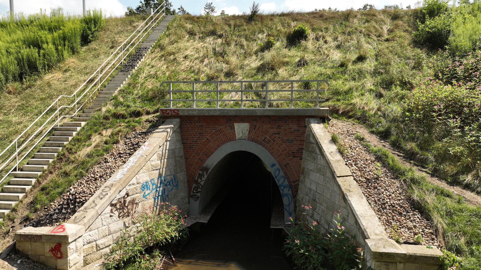 Ilustracja przedstawia przepust. Jest on niskim i wąskim tunelem wbudowanym w nasyp kolejowy, przez który przepływa strumień. Po lewej stronie zdjęcia widać schody prowadzące na szczyt nasypu.