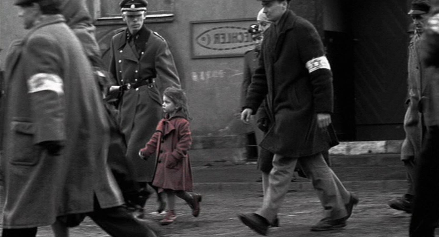 Zdjęcie czarno-białe przedstawia ulicę. Idą nią ludzie z opaskami z gwiazdą Dawida eskortowani przez Niemców. Pomiędzy nimi idzie dziewczynka w czerwonym płaszczyku.