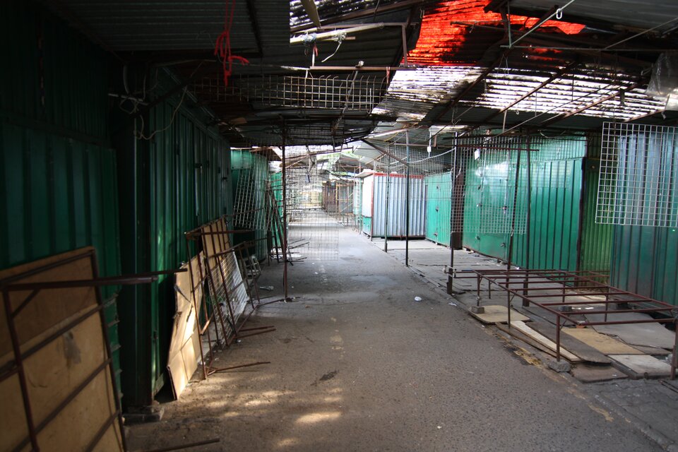 Zdjęcie przedstawia opuszczone blaszane boksy handlowe ustawione wzdłuż asfaltowej alejki, oddzielone metalową siatką. Na ziemi widać porozrzucane, puste kartonowe pudełka.