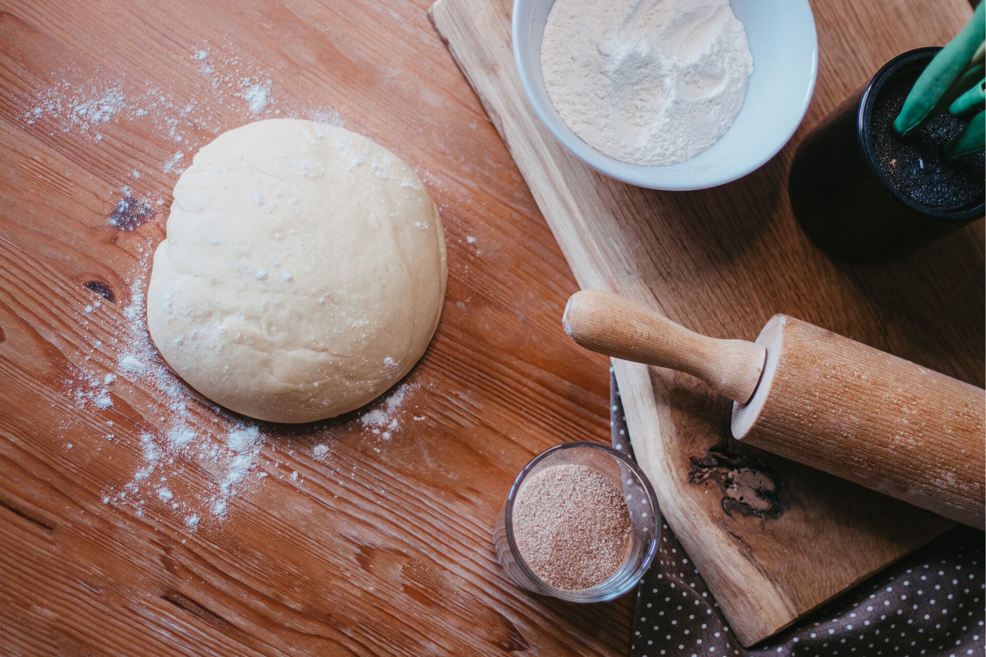 Zdjęcie przedstawia krok pierwszy formowania serduszek. Ujęcie wykonane jest od góry pod skosem. W lewej części zdjęcia widoczna jest jasna kula ciasta oprószona mąką. Leży ona na ciemnobrązowym drewnianym blacie. Dookoła ciasta rozsypana jest biała mąka. W prawym dolnym rogu dostrzec można ciemną ścierkę w białe kropki. Po jej lewej stronie widoczna jest od góry przezroczysta szklanka wypełniona do połowy cukrem cynamonowy. W prawej części zdjęcia znajduje się duża drewniana deska. Na desce tej w prawym dolnym rogu leży drewniany wałek, a obok po lewej stronie widoczny jest czarny owalny stojak w którym znajdują się noże z zieloną rączką. W górnym prawym rogu na drewnianej desce stoi biała miseczka widoczna od góry, wypełniona białą mąką.