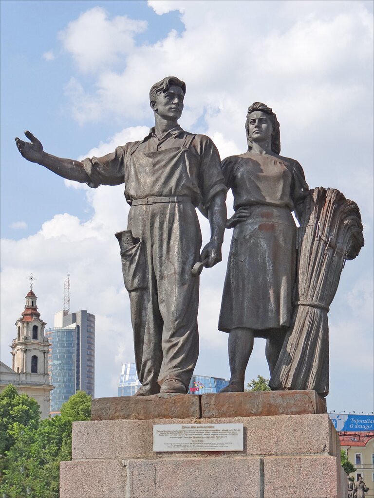 Zdjęcie przedstawia pomnik robotnika i kobiety pracującej na roli. Stoją obok siebie. Robotnik ma na sobie fartuch, prawą rękę ma uniesioną, w lewej trzyma narzędzie. Kobieta ma prostą suknię, prawą rękę opiera na biodrze, a w lewej trzyma snopek zboża.