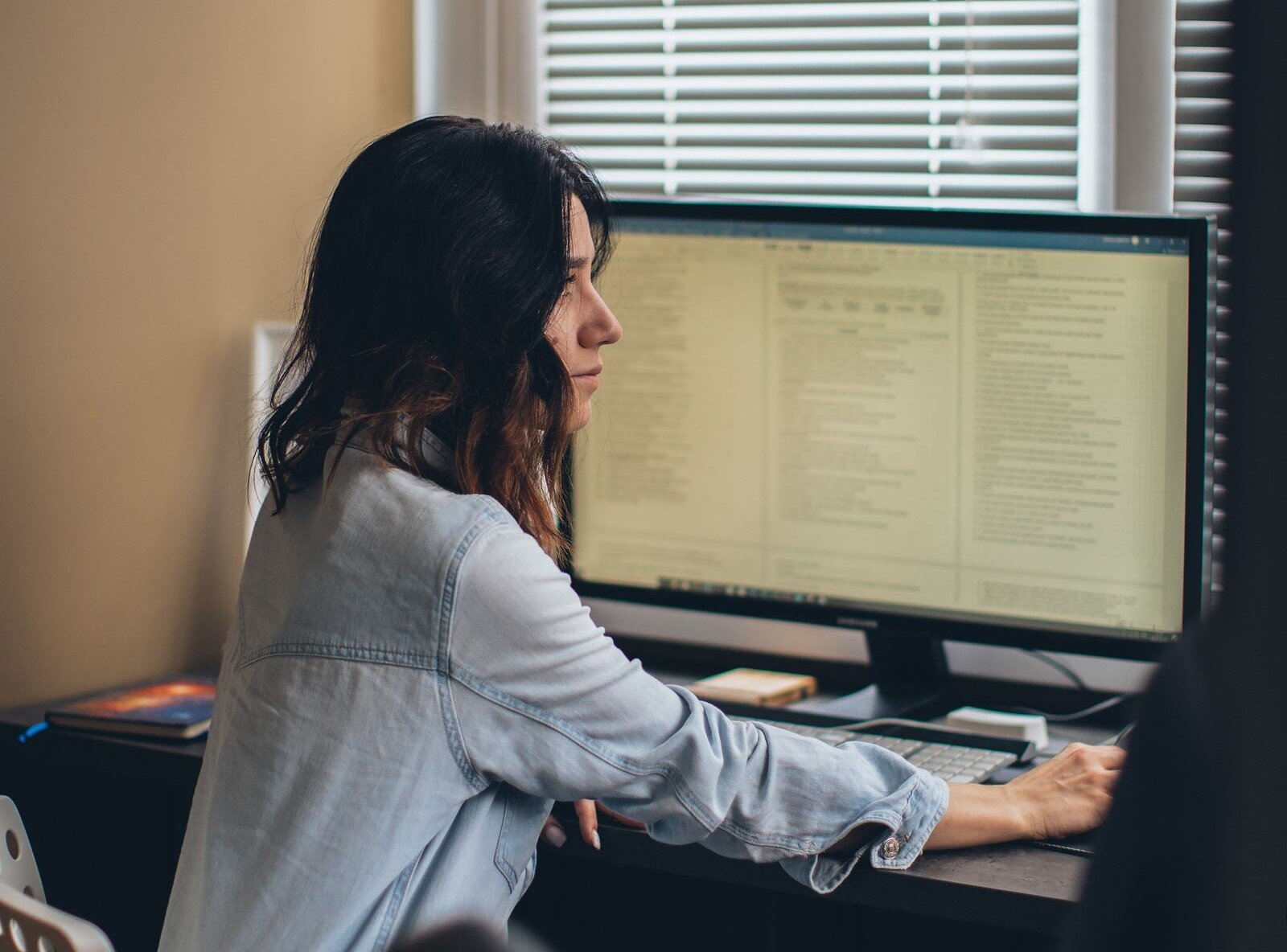 Zdjęcie przedstawia młodą kobietę od pasa w górę, siedzącą przy biurku i zwróconą w prawą stronę. Kobieta ubrana jest w dżinsową koszulę z długim rękawem, ma dłuższe i ciemne włosy. Jej prawa ręka leży na biurku, na którym stoi duży monitor komputera. Na ekranie komputera widoczne są otwarte dokumenty z tekstem. Przed monitorem widoczna jest klawiatura, a obok karteczki do robienia notatek. W tle widoczne jest okno zasłonięte żaluzjami.