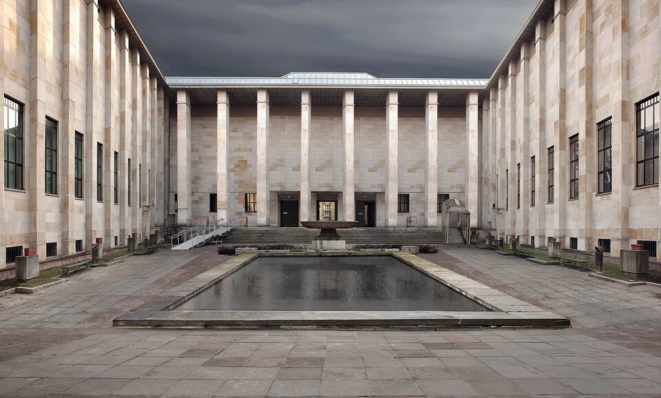 Zdjęcie przedstawia dziedziniec Muzeum Narodowego w Warszawie. Pośrodku dziedzińca znajduje się prostokątny, płytki zbiornik z wodą. Jest on otoczony ścianami budynku.