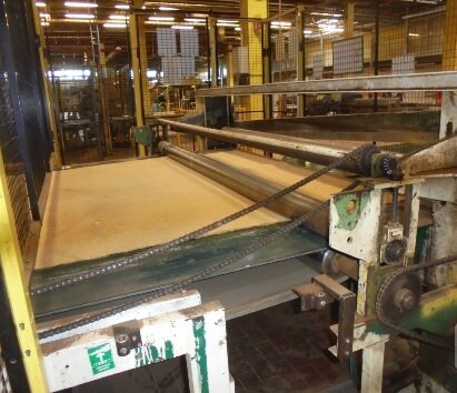 Zdjęcie przedstawia gilotynę - maszynę do produkcji wyrobów z włókna kokosowego. Stoi w hali zakładu produkcyjnego. Po bokach i z tyłu jest zabezpieczona ogrodzeniem z siatki. Po lewej stronie taśma z włóknem kokosowym, które wprowadzane jest pomiędzy wałki. Przed taśmą widoczne łańcuchy napędowe na kołach zębatych.