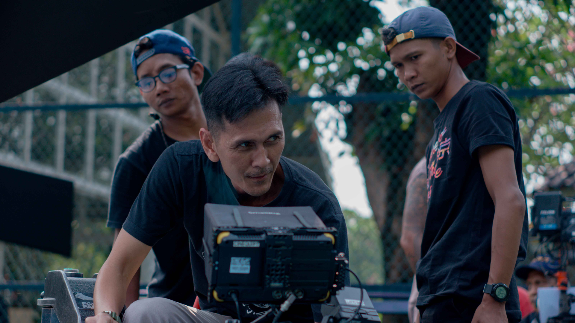 Zdjęcie przedstawia mężczyznę/reżysera, który spogląda na ekran kamery stojącej przed nim. Za reżyserem znajduje się dwóch młodych mężczyzn. Scena rozgrywa się na powietrzu.