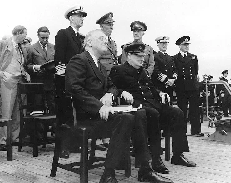 Kliknij, aby powiększyć Zdjęcie przedstawia dwóch mężczyzn - prezydent USA Franklina D. Roosevelta i premiera Wielkiej Brytanii Winstona Churchilla. Siedzą na krześle. Jeden z nich ubrany jest w garnitur i nosi okulary. Drugi ubrany jest w mundur galowy. Za ich plecami stoją umundurowani oficerowie i mężczyźni w marynarkach.