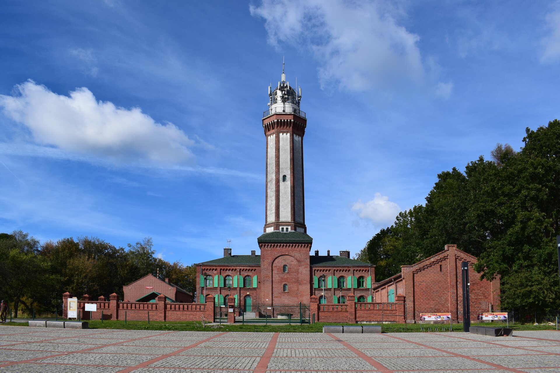 Fotografia latarni morskiej. U podnóża kilkunastometrowej wieży latarni znajduje się jednopiętrowy budynek z czerwonej cegły z zielonymi okiennicami. Na wieży taras widokowy i zamontowane anteny. Zwieńczona niewielką kopułą. 