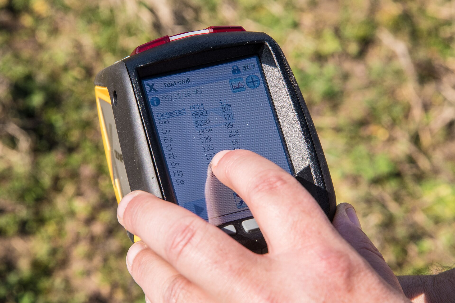 Zdjęcie przedstawia urządzenie niewielkich rozmiarów trzymane w dłoni. Urządzenie ma wyświetlacz z danymi. Widać symbole pierwiastków oraz wartości liczbowe. W tle urządzenia jest powierzchnia ziemi porośnięta niską roślinnością.  