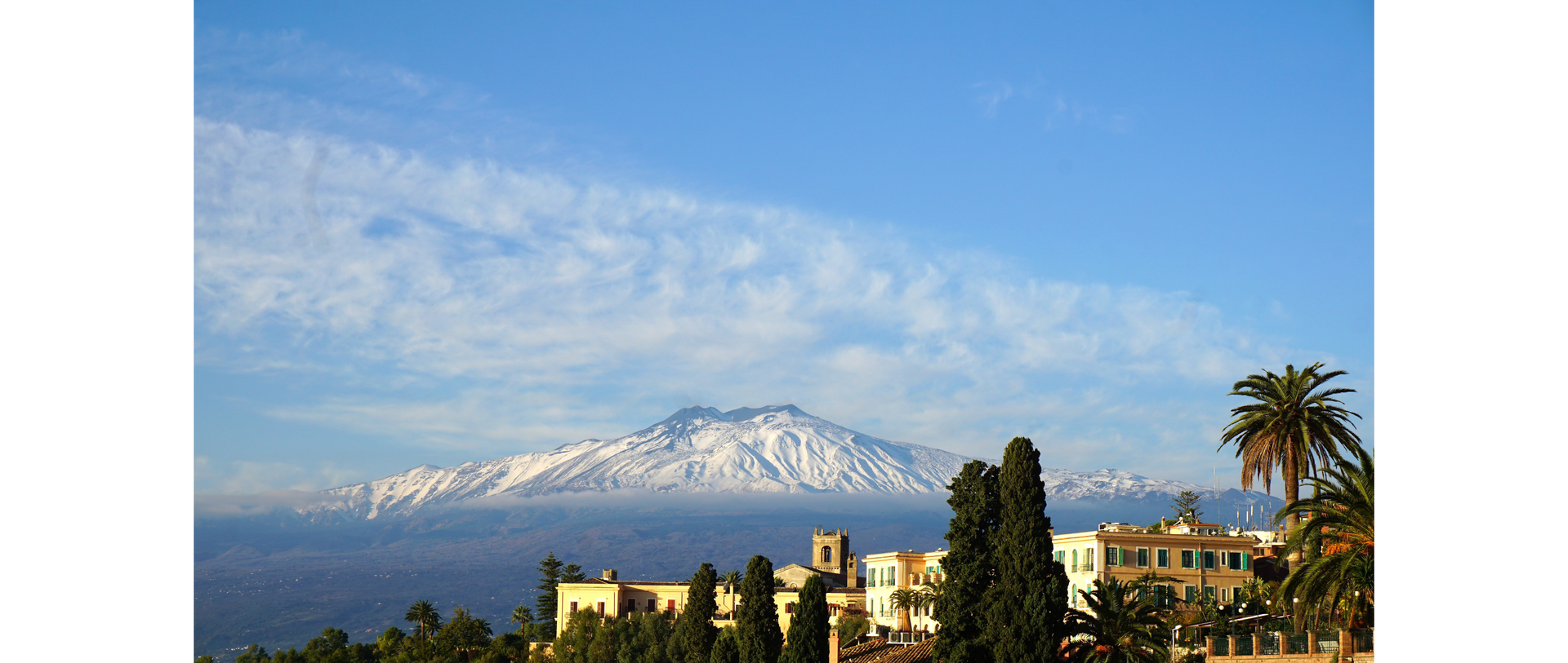 Zdjęcie przedstawia słynny sycylijski wulkan Etnę. Zdjęcie zostało wykonane z punktu widokowego we włoskim miasteczku Taormina. Na zdjęciu widoczne są fragmenty budynków, dachy oraz roślinność.