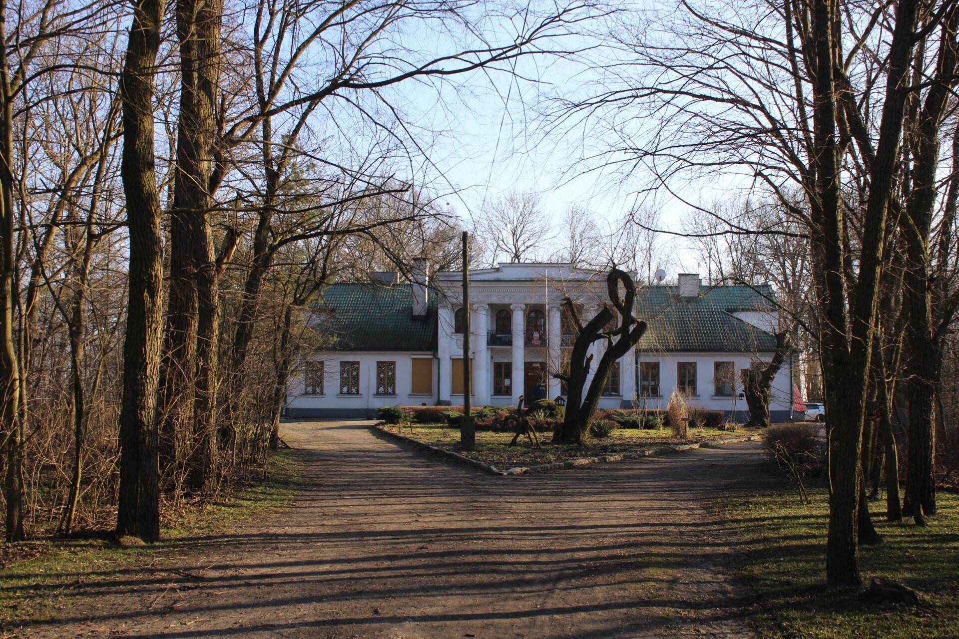 Zdjęcie przedstawia biały dworek z szarym dachem. Środkowa część budynku jest lekko wysunięta do przodu i wsparta kolumnami. Okna rozmieszczone są symetrycznie, część z nich jest zakryta brązową dyktą. Do dworku prowadzi piaskowa droga. Z obu stron drogi, przed dworkiem, rosną wysokie, bezlistne drzewa. Niebo jest błękitne, bezchmurne. Zdjęcie zostało wykonane w świetle dziennym.