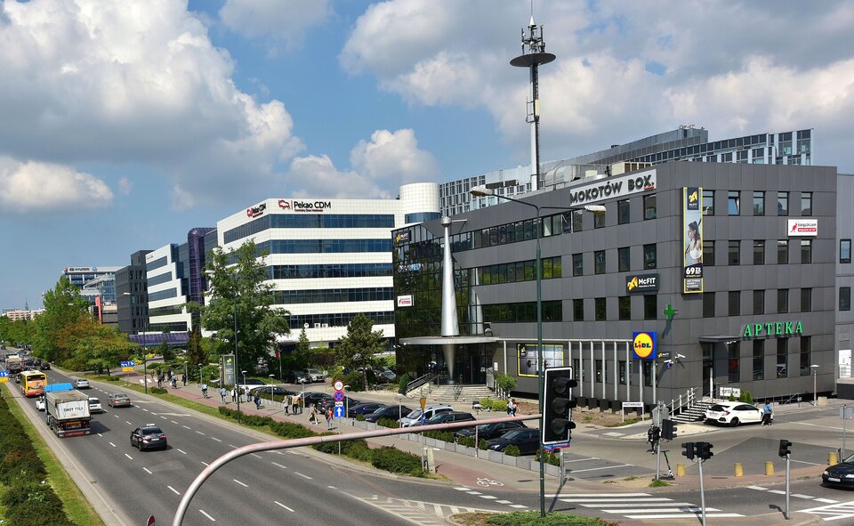 Zdjęcie przedstawia biurowce stojące przy ruchliwej drodze. Budynki biurowców, między innymi Pekao i Mokotów Box., są kilkukondygnacyjne. Mają liczne okna.  Przy budynkach są parkingi.  