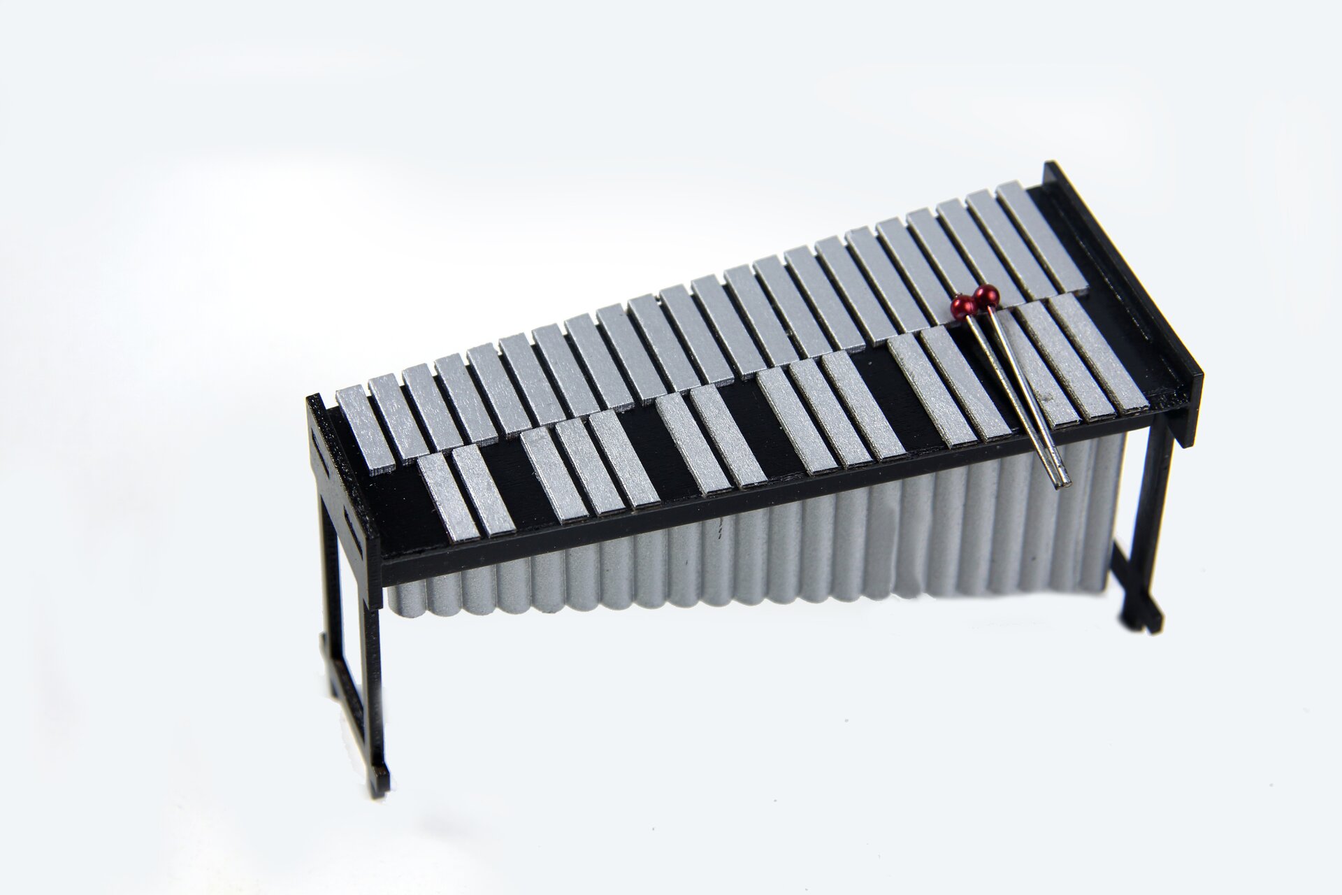 Fotografia przedstawia wibrafon. Instrument perkusyjny zbudowany z czarnego stelażu, na którym rozmieszczone zostały srebrne płytki pod którymi wiszą tuby. Na instrumencie leżą dwie pałeczki.